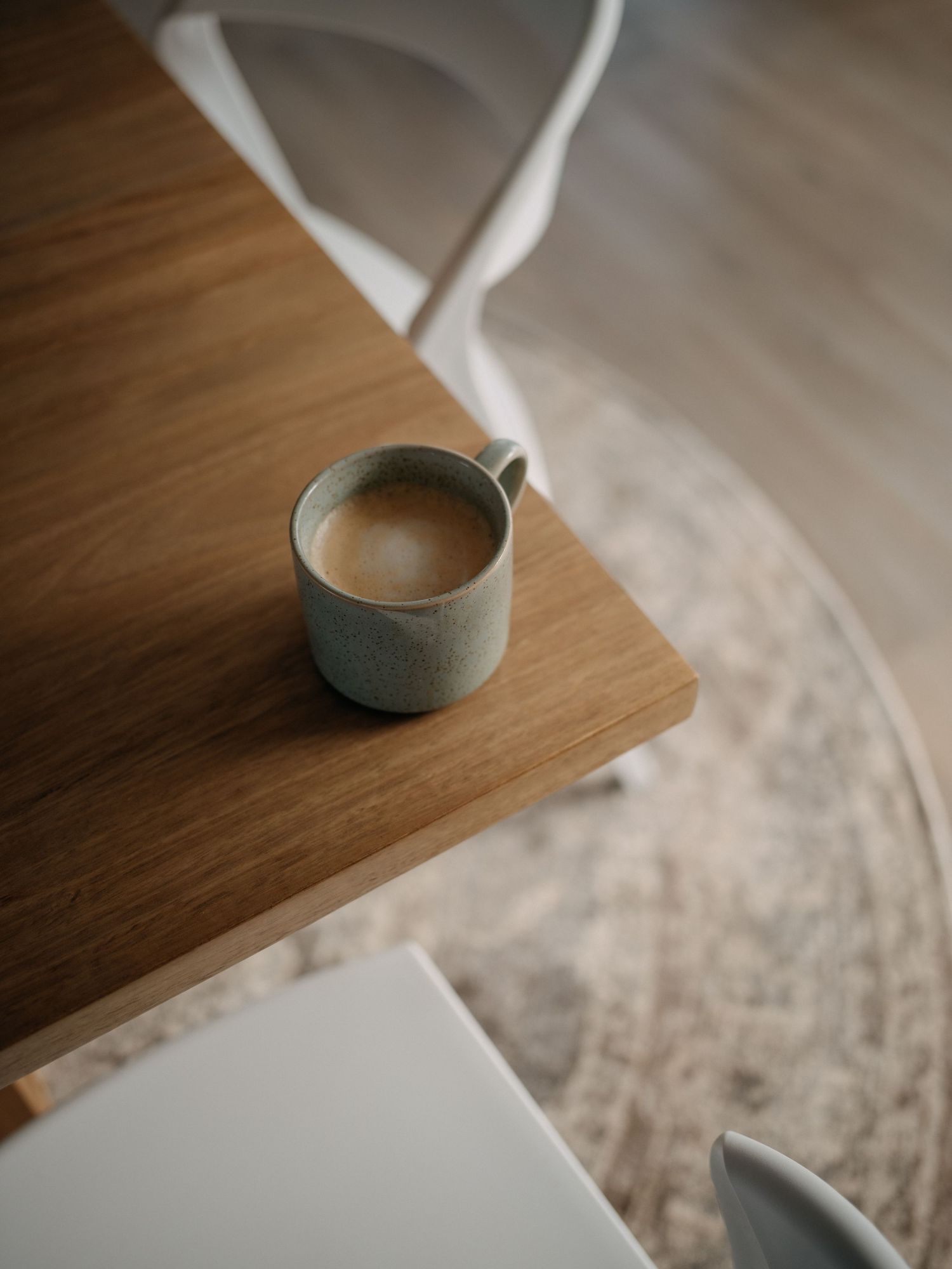 A ceramic mug of coffee sits on a wooden table beside a white chair on a neutral rug.
