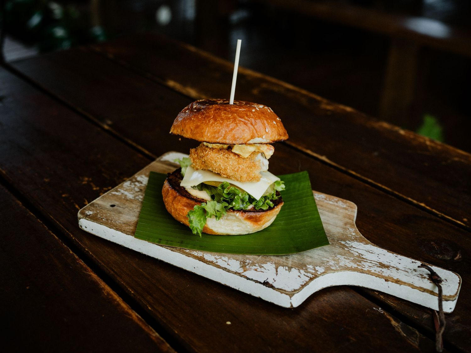 A gourmet burger with fresh lettuce and toppings on a rustic white wooden board with a banana leaf.