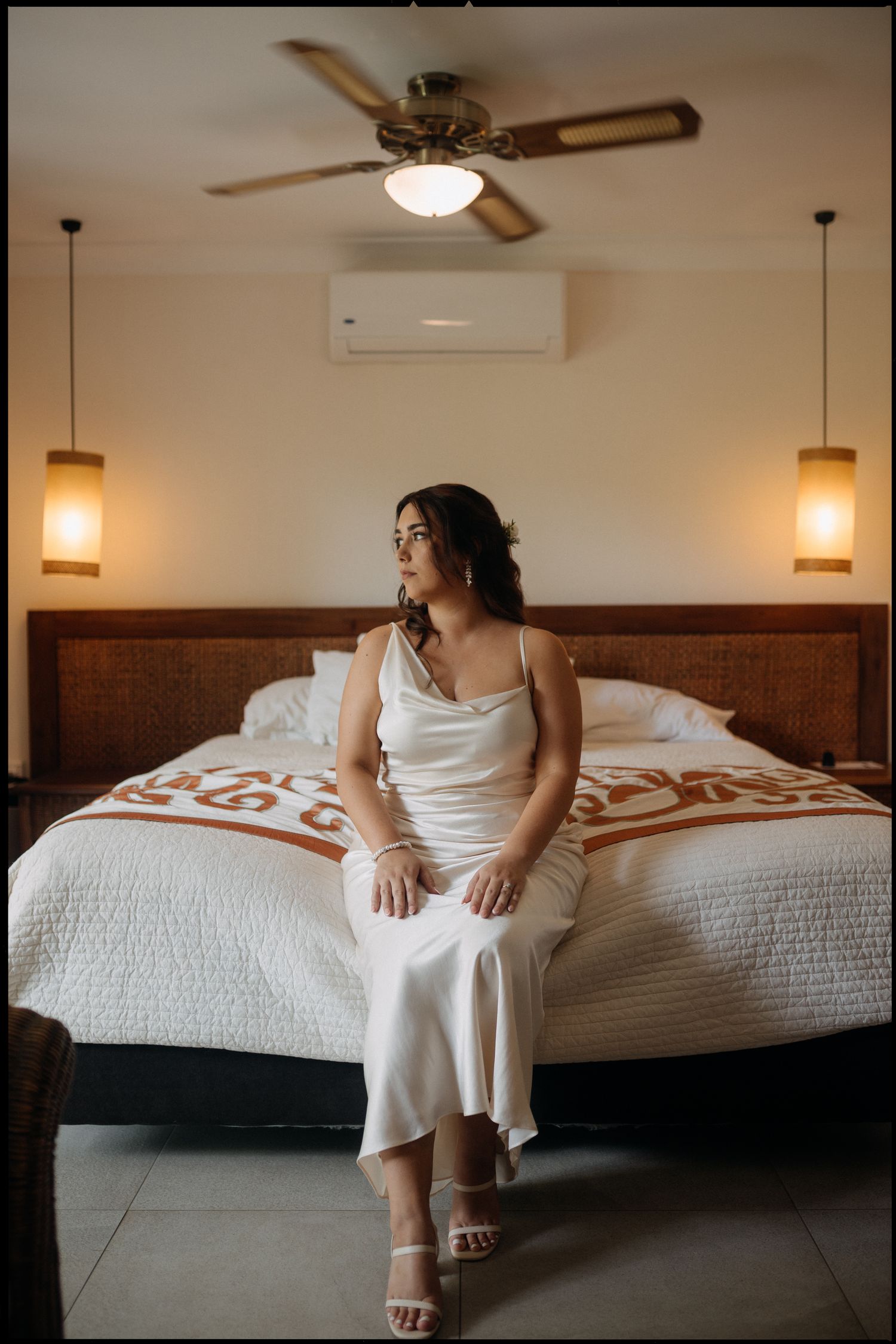 Bride in white satin slip dress and heels sitting on a hotel bed with ceiling fan and warm pendant lights.