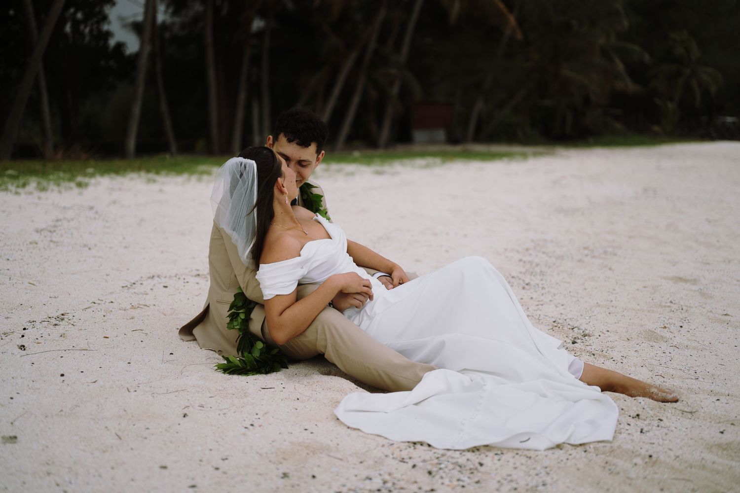Couple in wedding attire relaxing on white sand beach, bride in white gown leaning against groom near tropical palm trees.