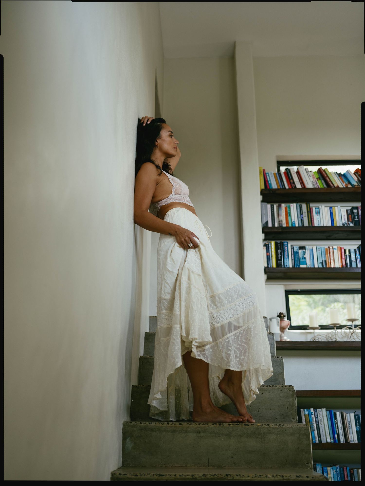 Woman in white skirt and crop top posing on stairs near a bookshelf in a modern interior.