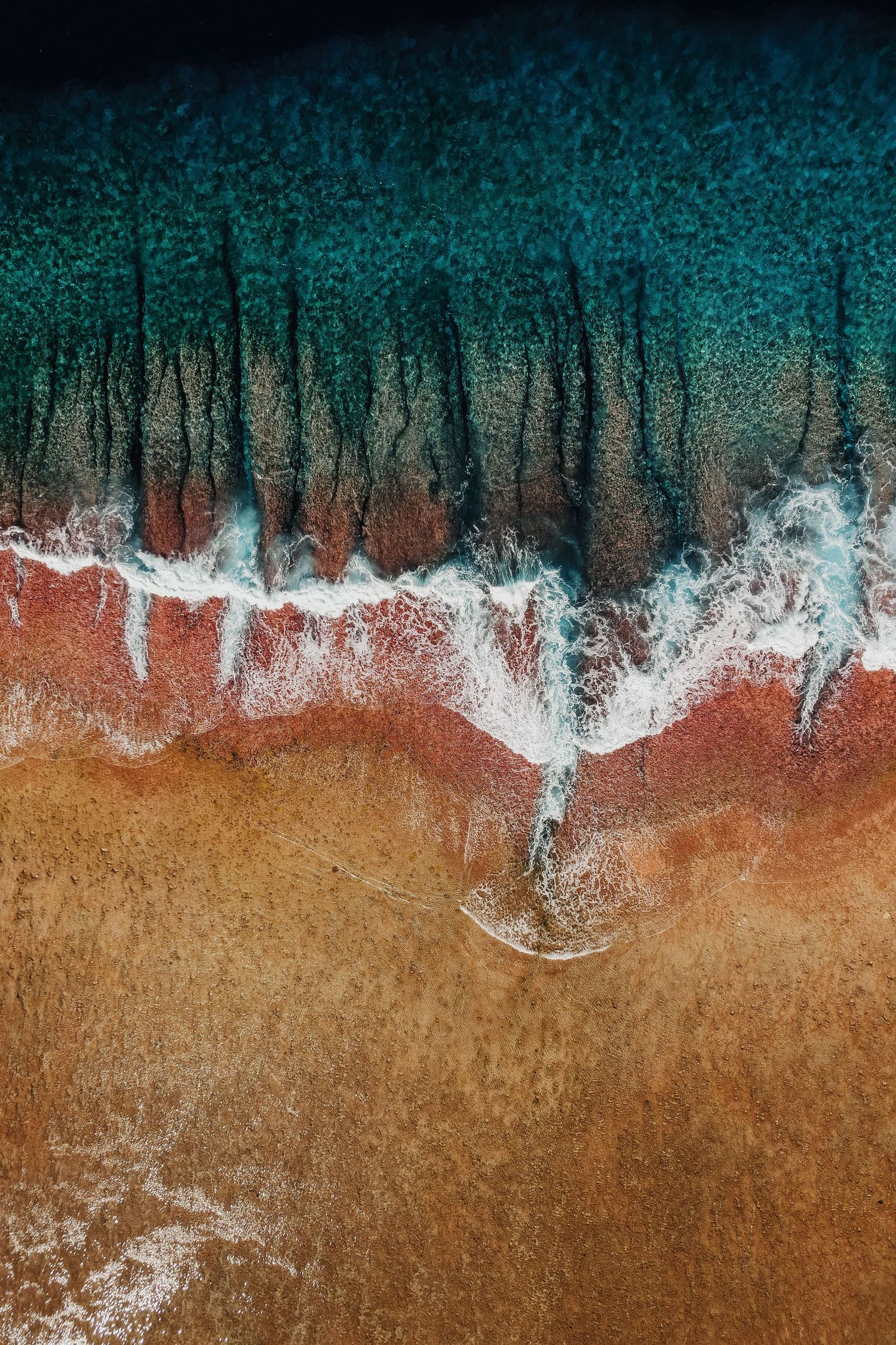 Aerial view of turquoise ocean waves washing onto a sandy beach with white foam and rich red-brown sand tones.