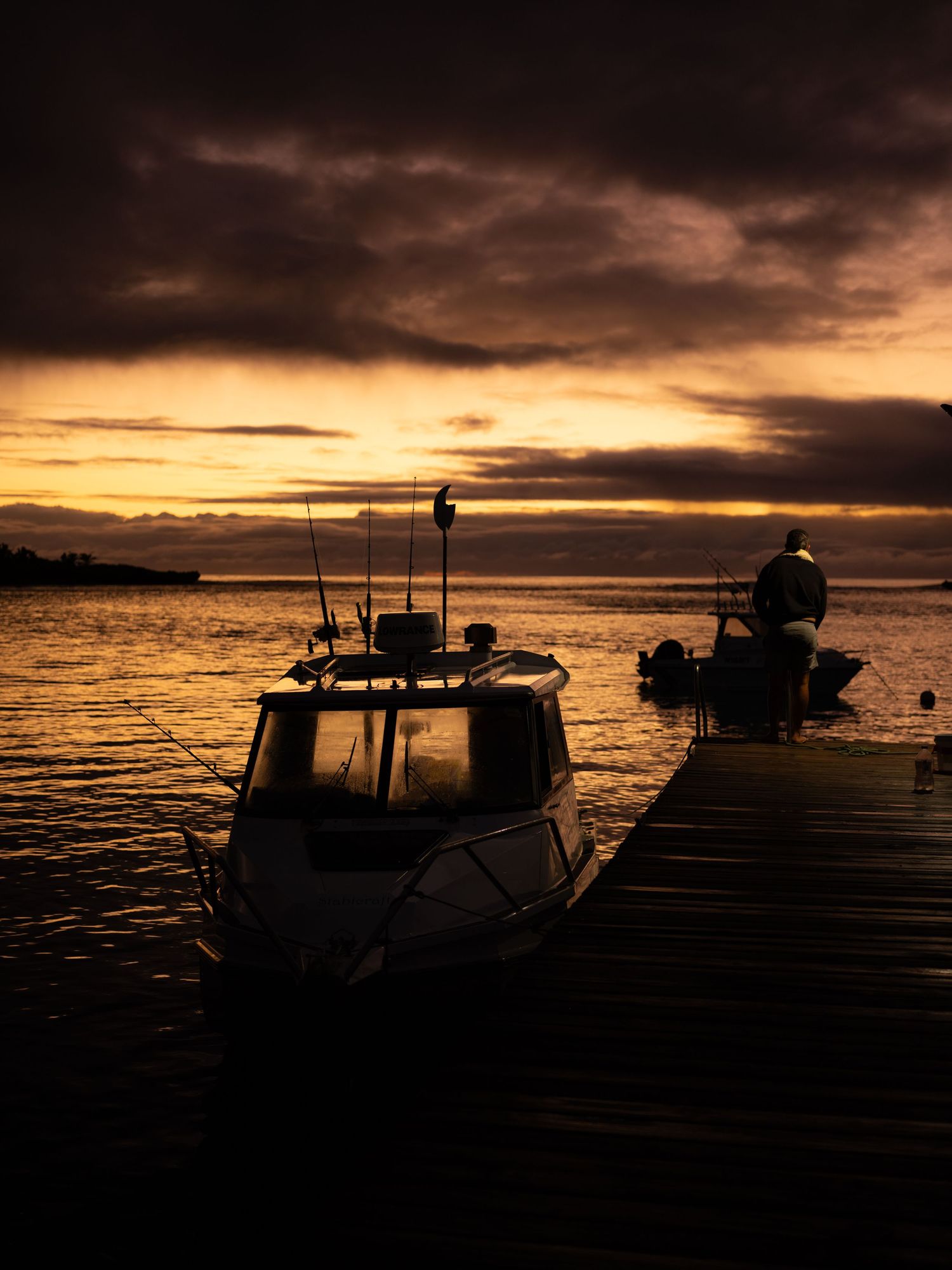 Silhouetted fishing boats moored at a wooden dock during a dramatic golden sunset over calm water.