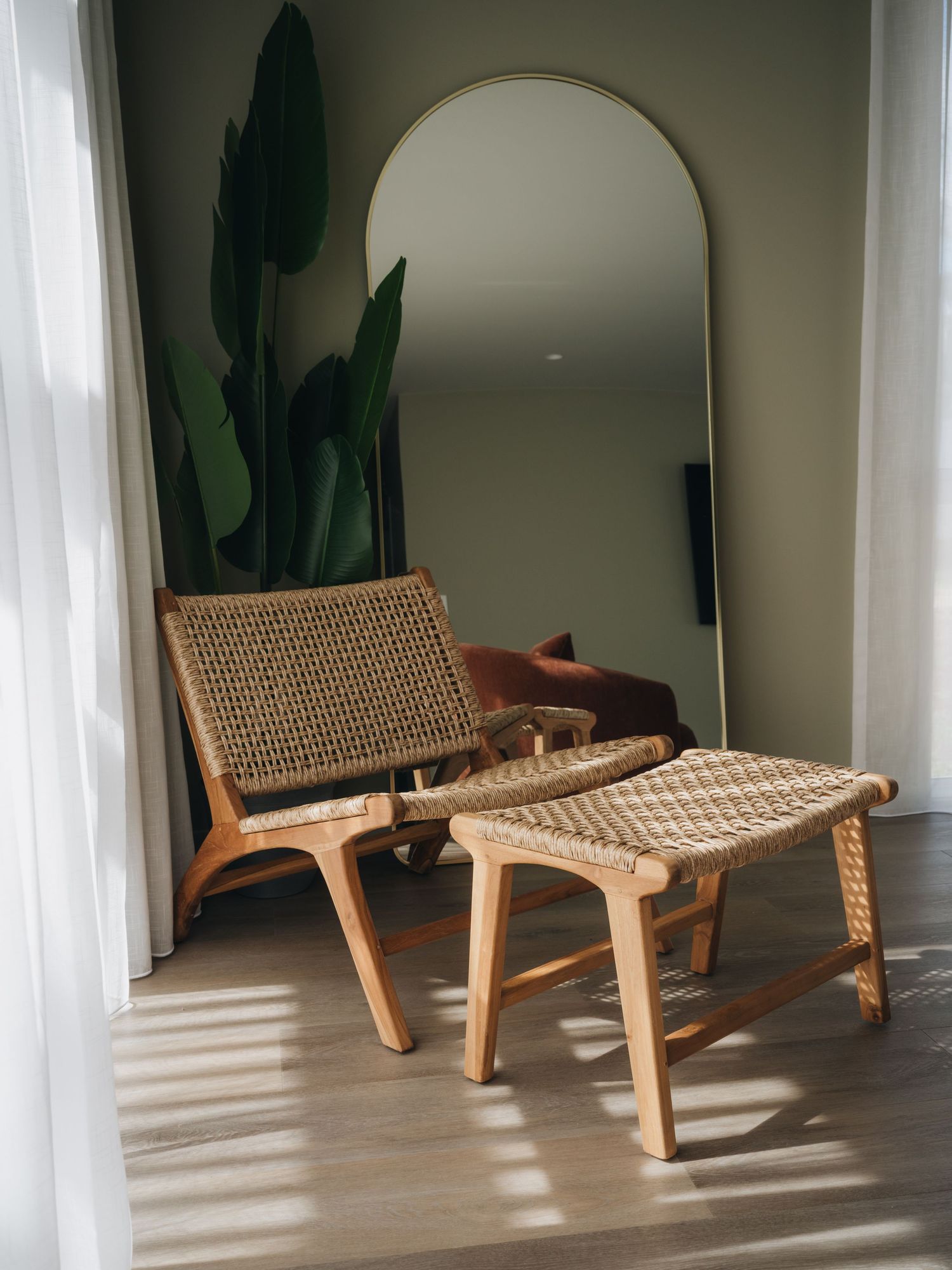 Woven rattan lounge chair with matching footstool beside a tall arch mirror on green wall with tropical plant.