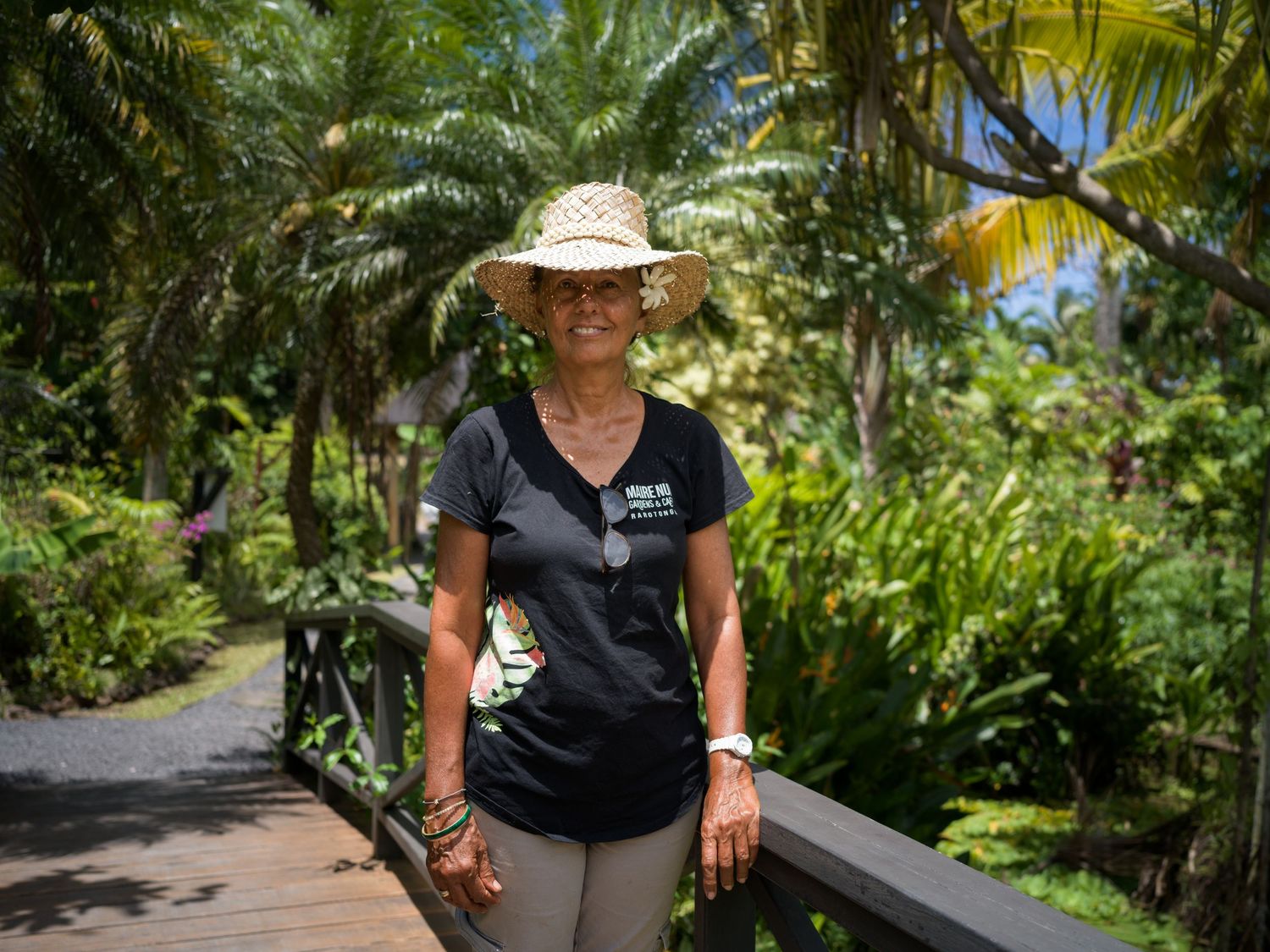 Woman in straw hat and black t-shirt smiling on a wooden bridge surrounded by lush tropical palm trees.