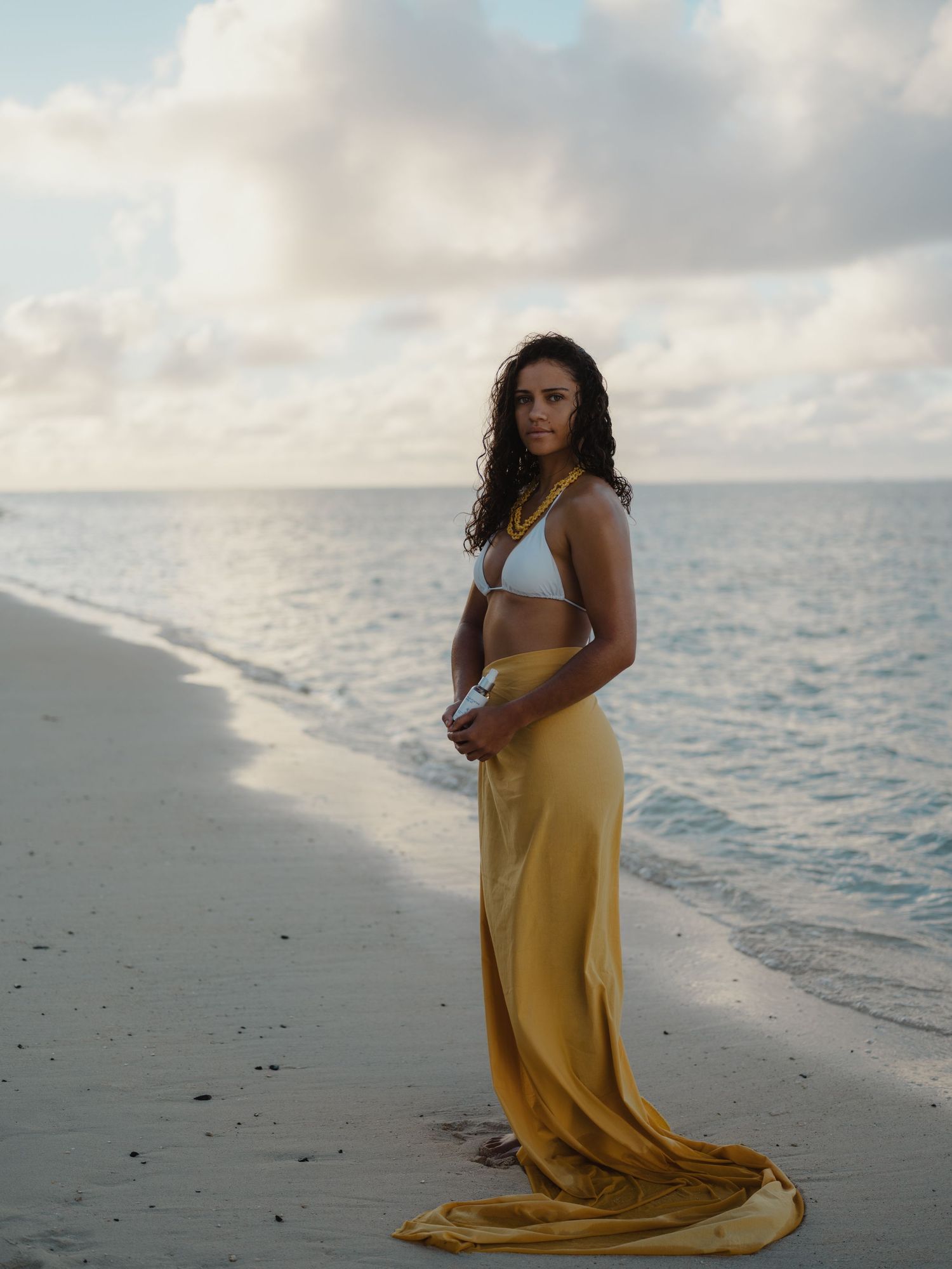 Woman in white bikini top and gold maxi skirt standing on a serene tropical beach at dusk.