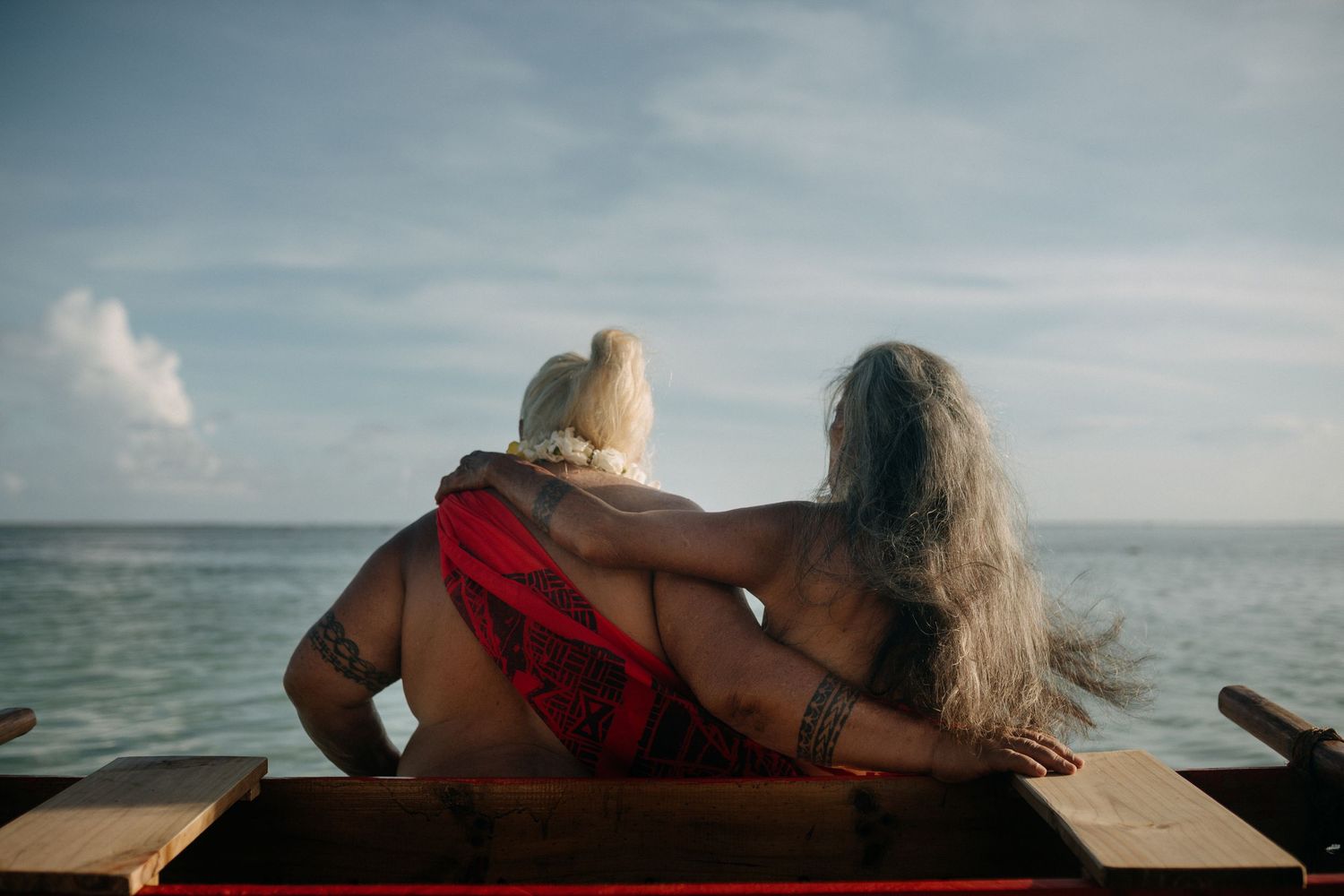 Two people with long hair sit embracing on a boat, gazing at a calm turquoise ocean under a partly cloudy sky.