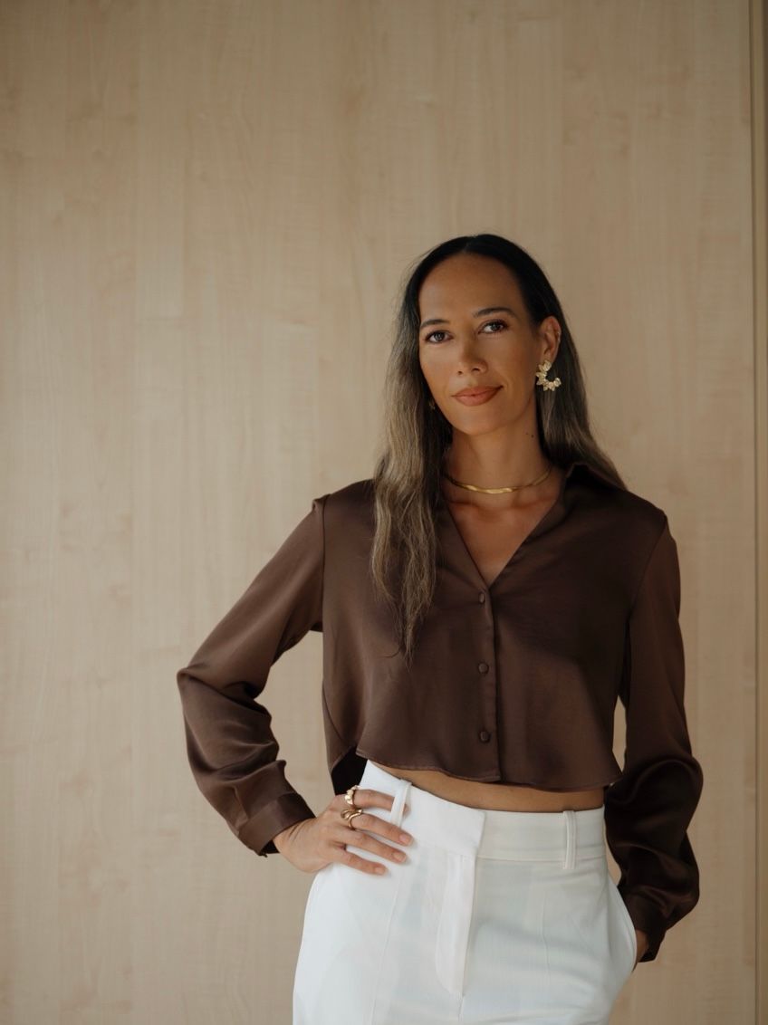 Woman in brown satin cropped blouse and white trousers with gold jewelry poses against wooden wall.