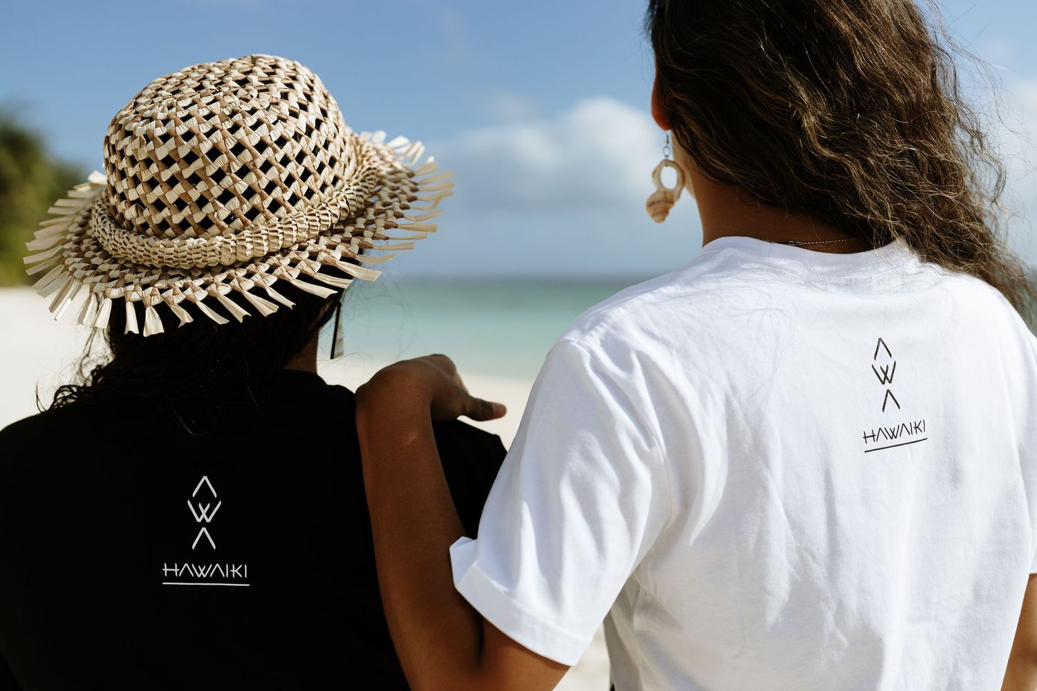 Two people wearing Hawaiki branded black and white t-shirts stand on a tropical beach, viewed from behind.