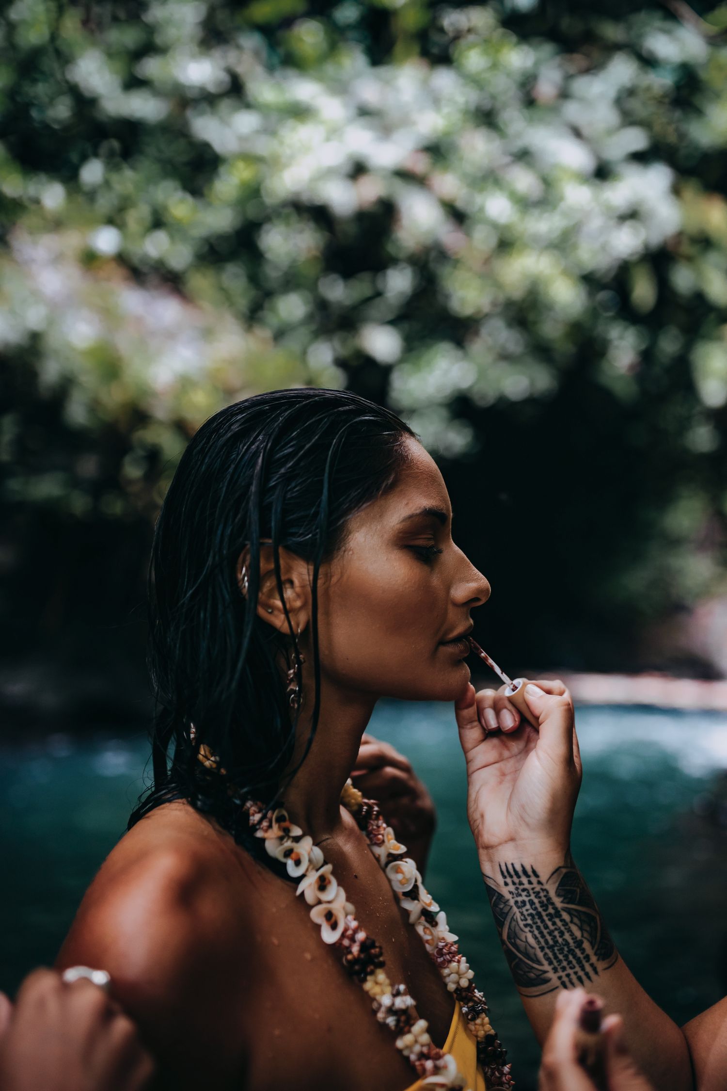 Woman with wet hair and floral lei applies lipstick outdoors near tropical greenery, showcasing tattoo and earrings.