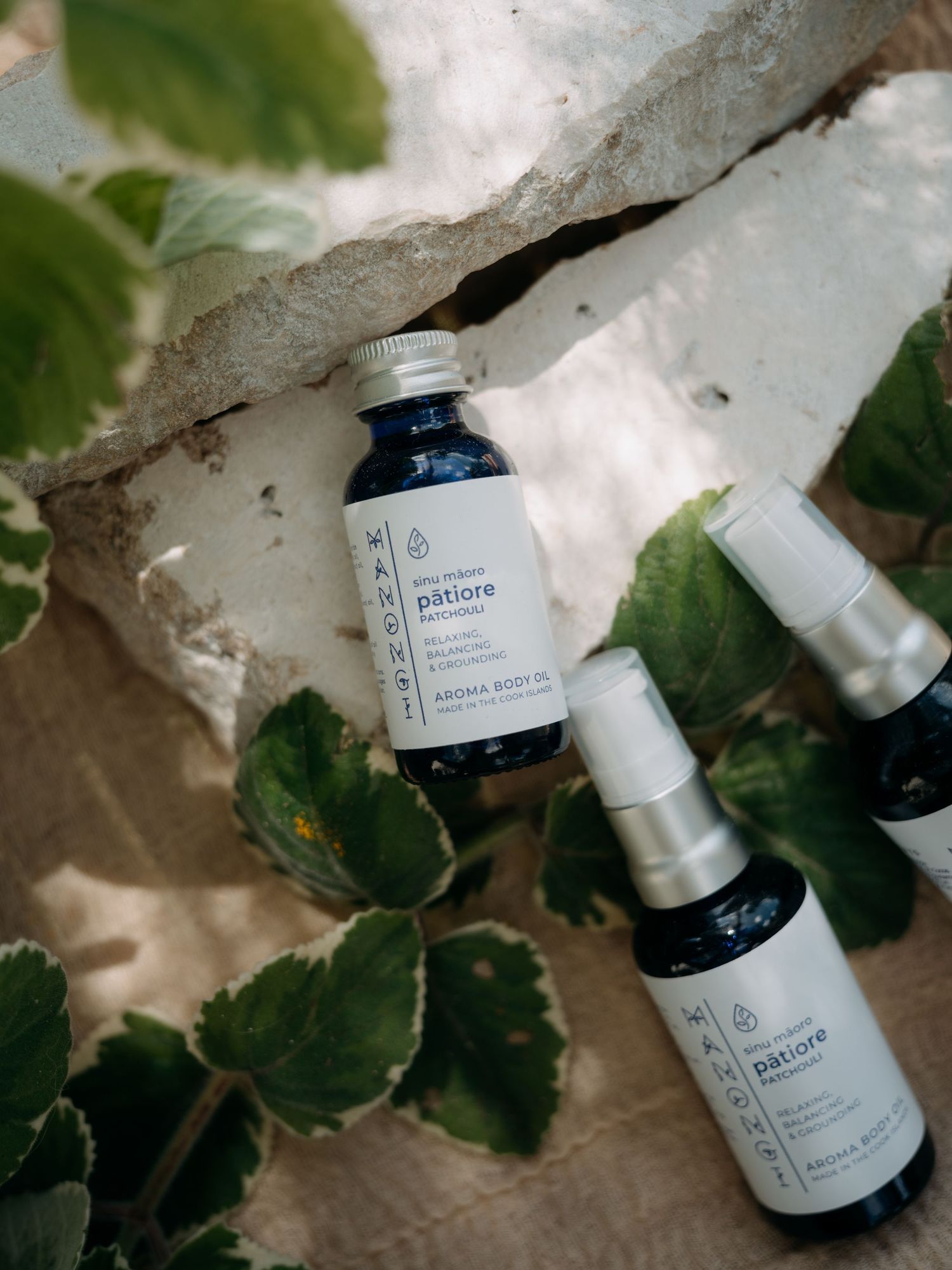 Patiore aroma body oil and spray bottles arranged on stone surface surrounded by lush green leaves.