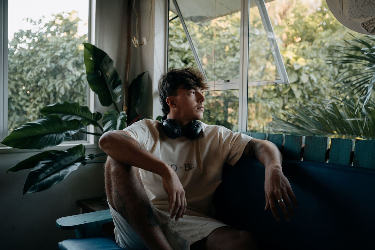Young man with headphones around neck sitting on couch near window surrounded by indoor plants in sunlit room.