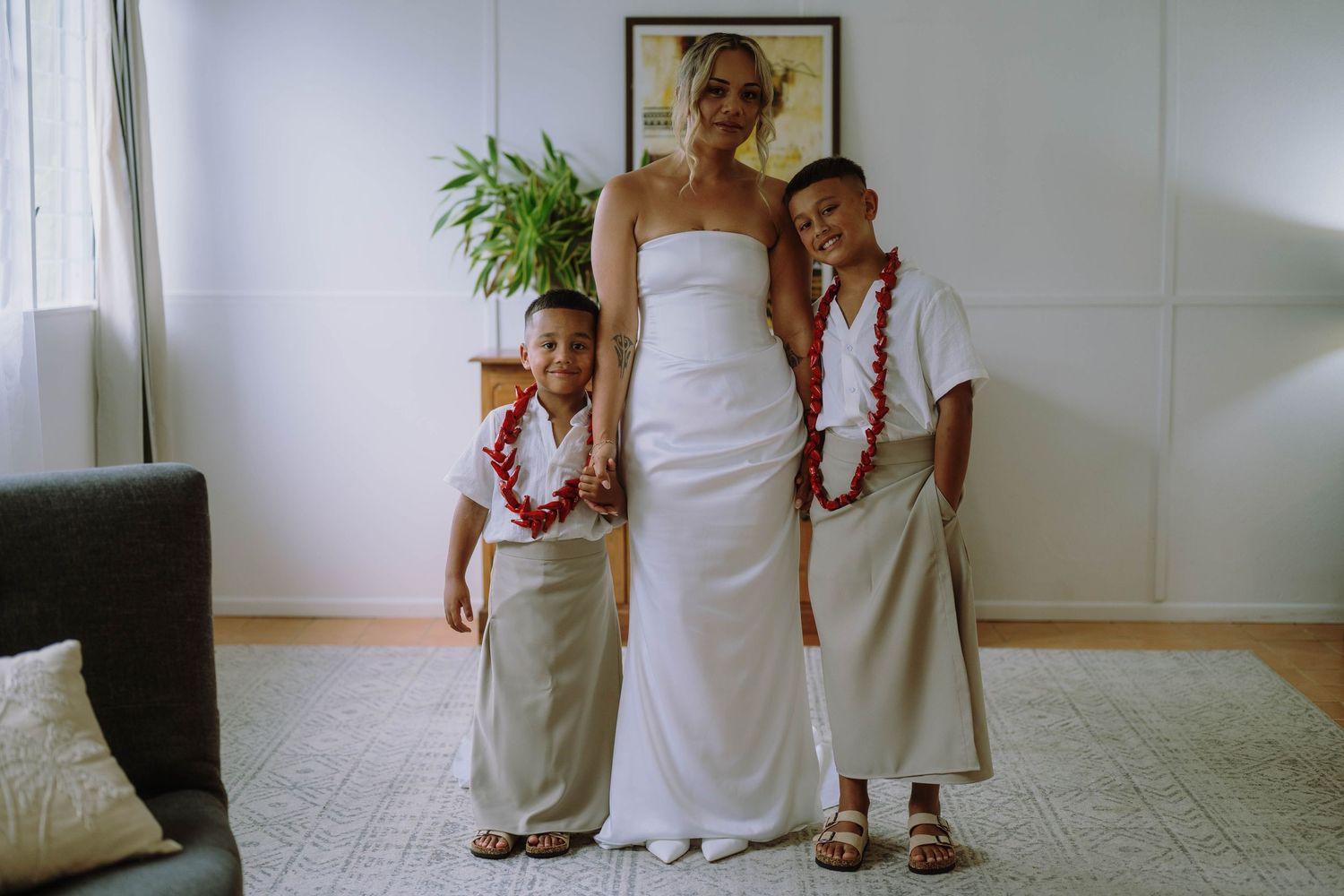 Bride in white strapless gown poses with two children wearing leis and traditional attire indoors.