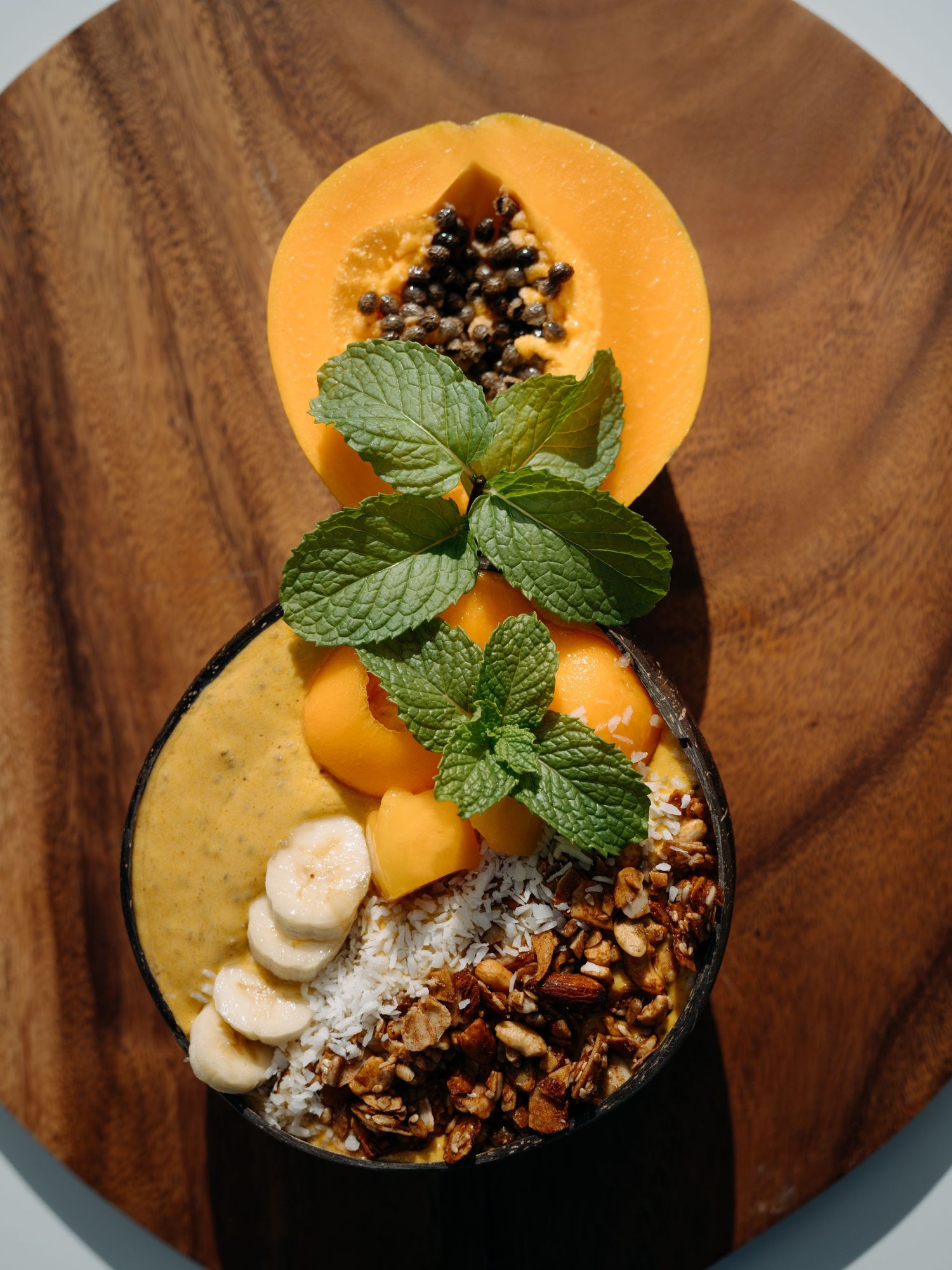 Tropical smoothie bowl with papaya, banana slices, granola, coconut, mango, and fresh mint on a wooden board.
