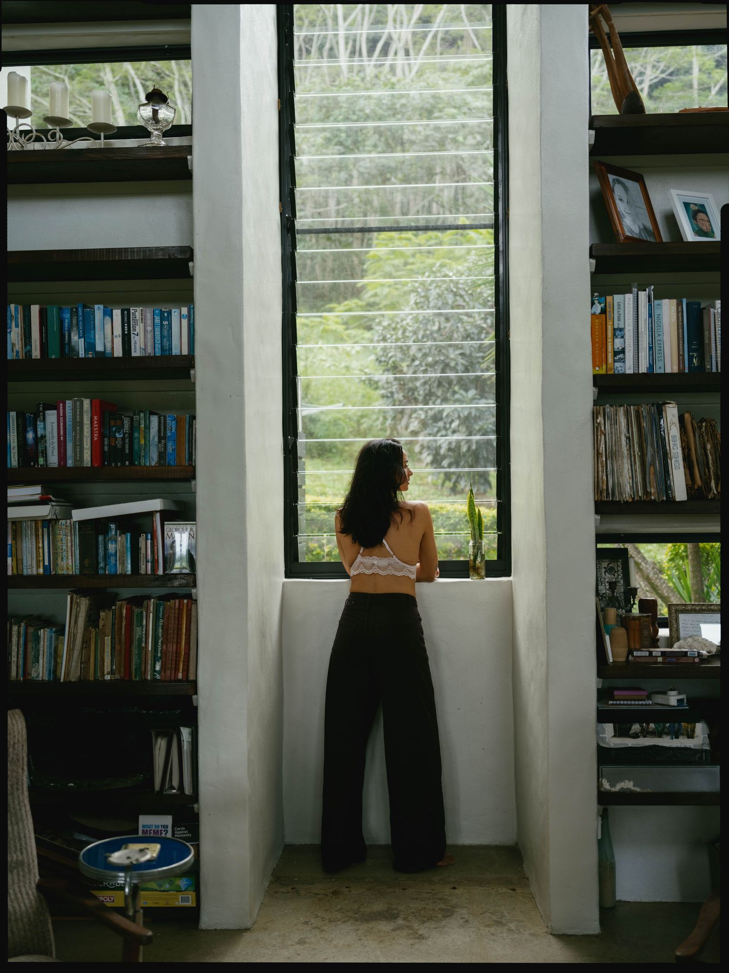 Woman standing between tall bookshelves gazing out a large window surrounded by lush green trees in a cozy library.