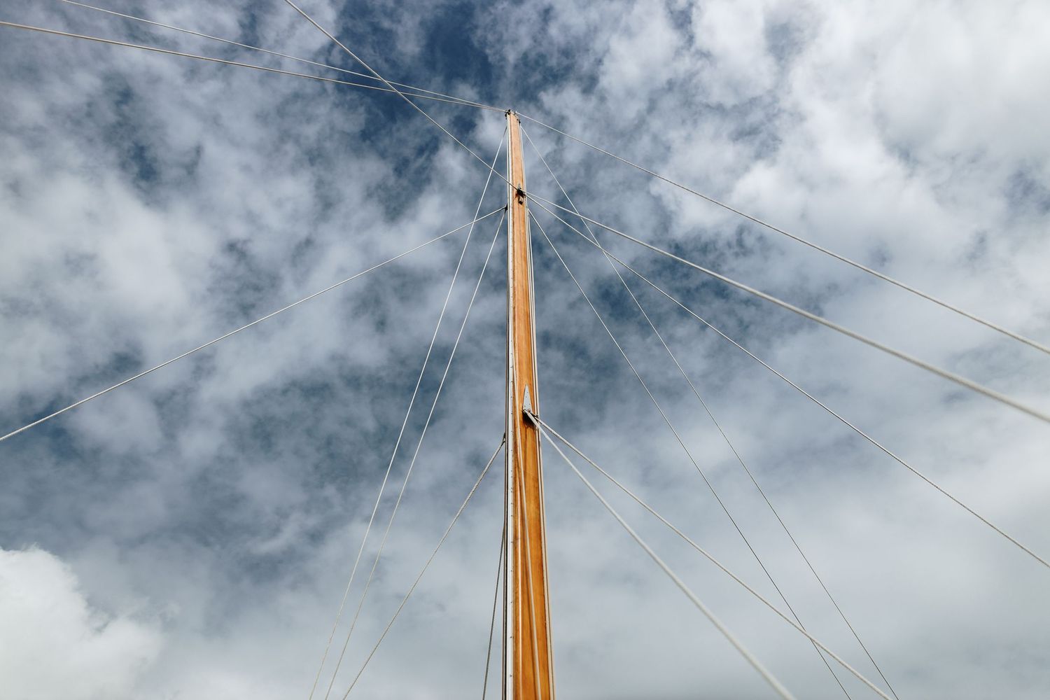 Tall wooden sailboat mast with wire rigging stretching against a cloudy blue sky, viewed from below.