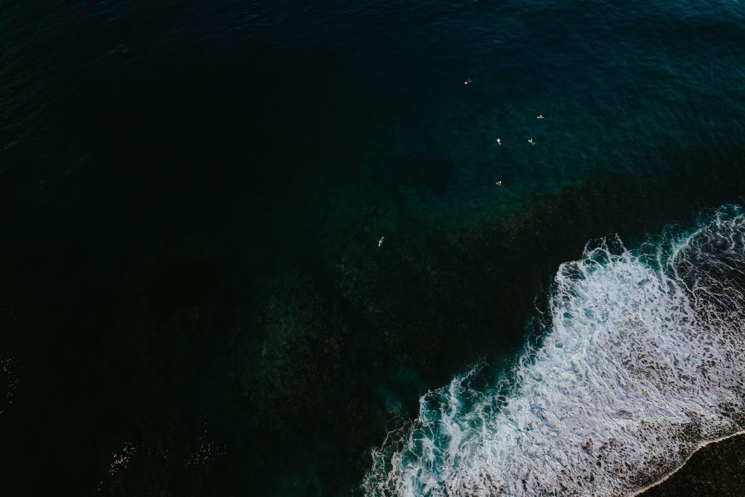 Aerial view of dark teal ocean waves crashing onto a sandy shore with surfers visible in the water.