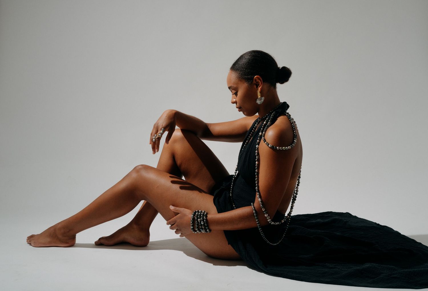 Woman wearing black draped fabric and beaded jewelry, seated gracefully on a white studio background.