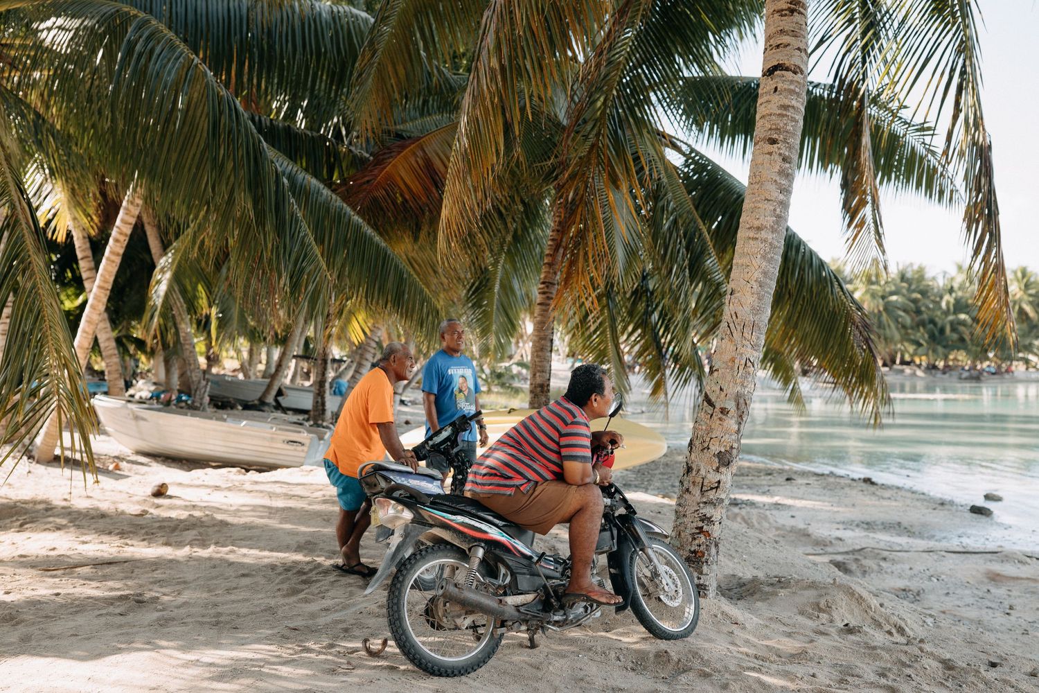 Men gathered around a motorcycle on a tropical beach shaded by palm trees with sandy shores in the background.