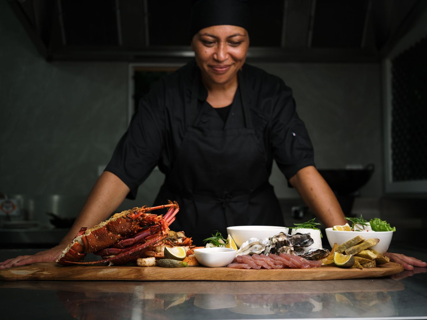 Smiling female chef presenting a large seafood platter with lobster, deli meats, and condiments in a restaurant kitchen.