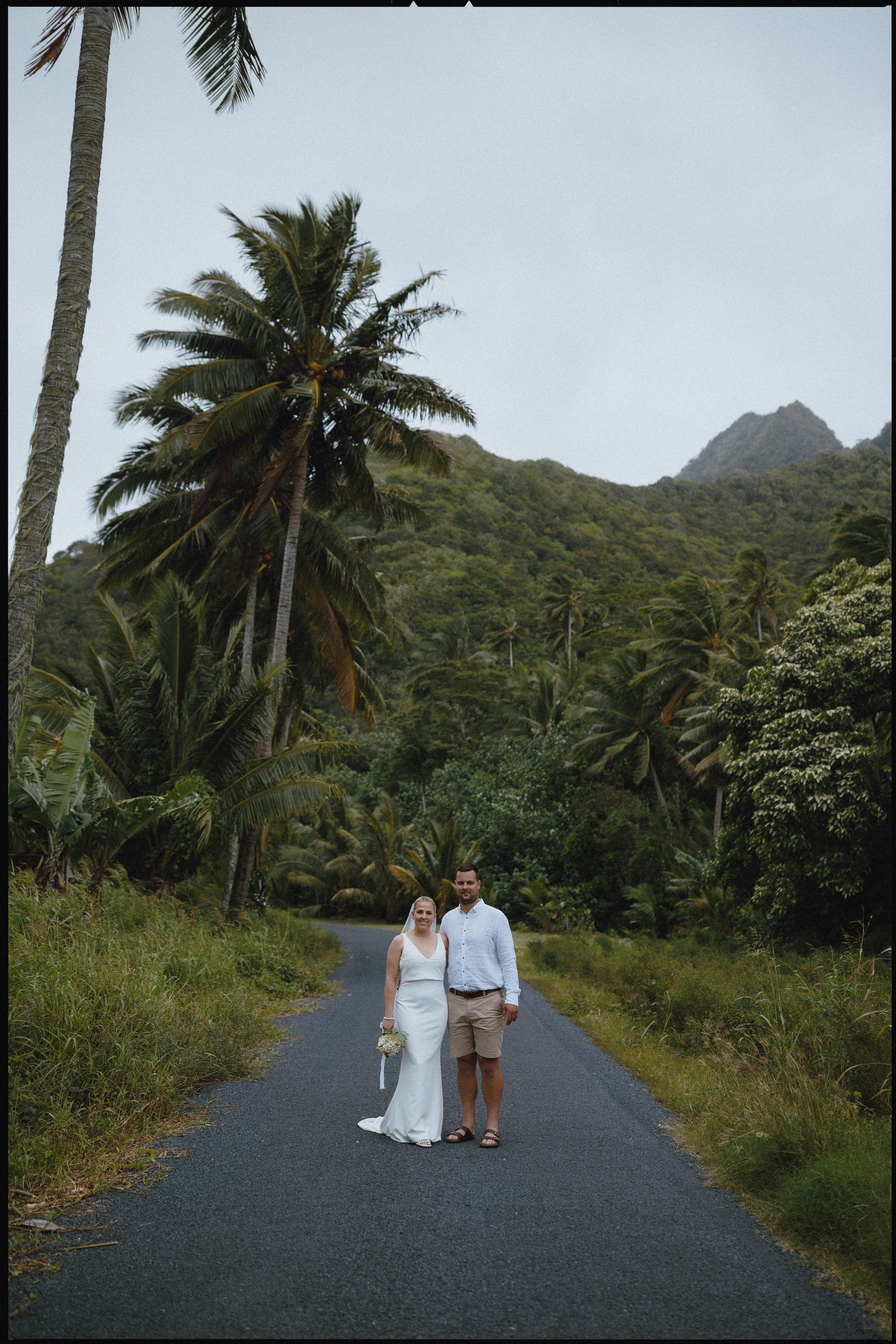 Newlywed couple posing on a tropical road surrounded by palm trees and lush mountains on their wedding day.