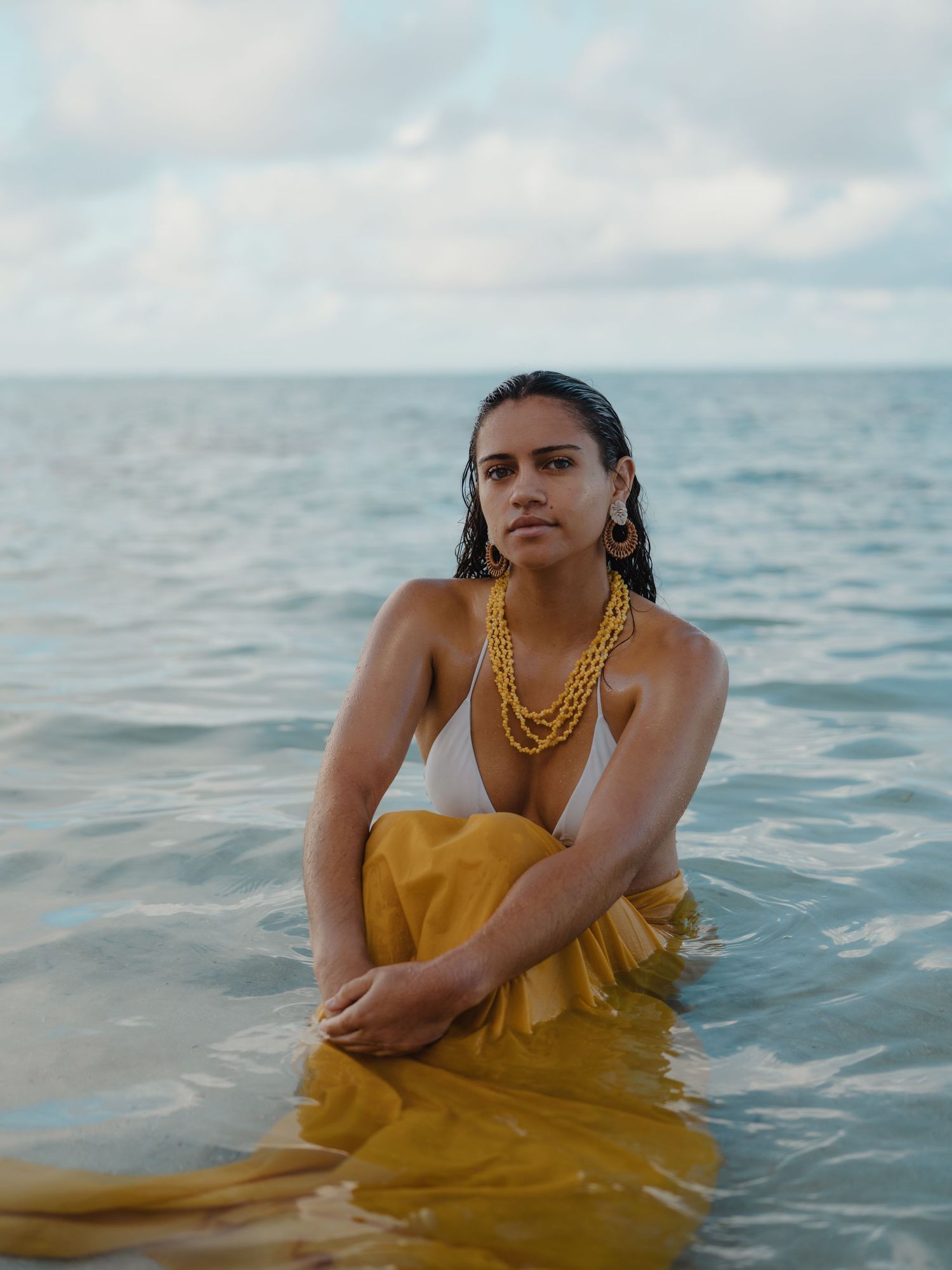 Woman in white bikini and yellow skirt wearing gold necklaces sitting in calm ocean waters under cloudy sky.