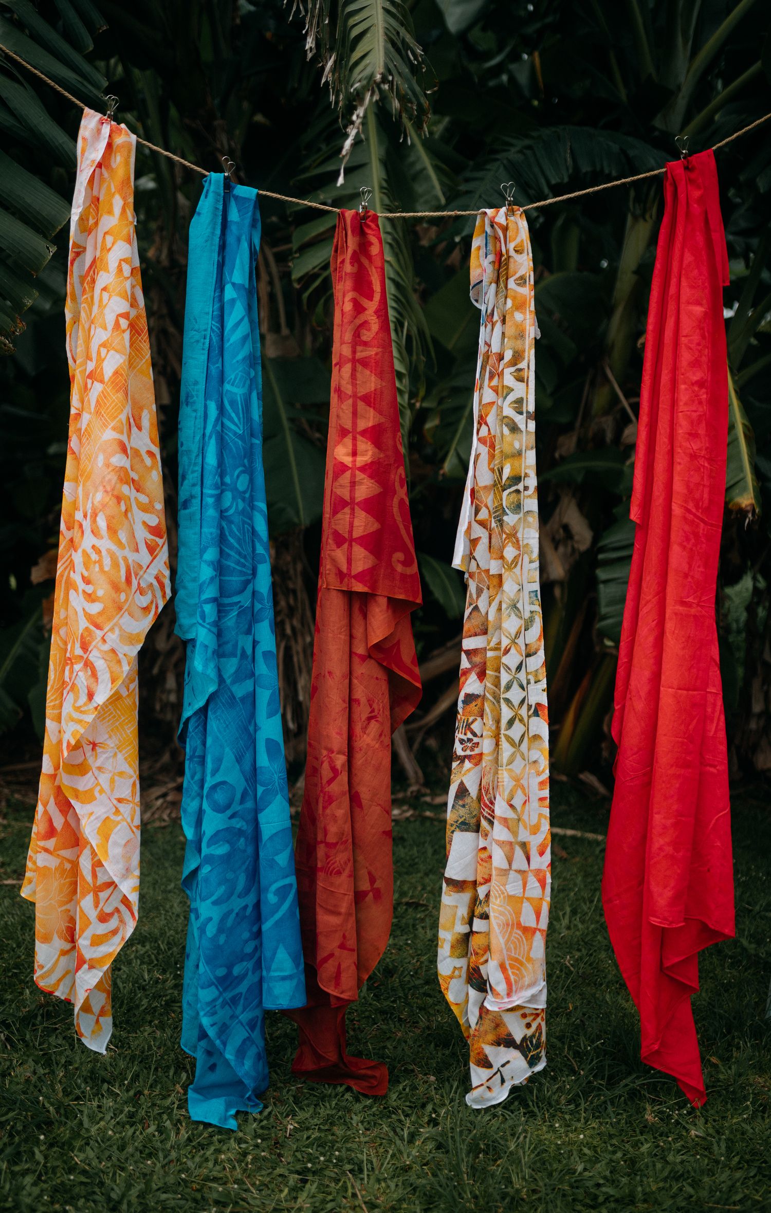 Colorful patterned fabric scarves hanging on a clothesline outdoors with tropical palm trees in the background.