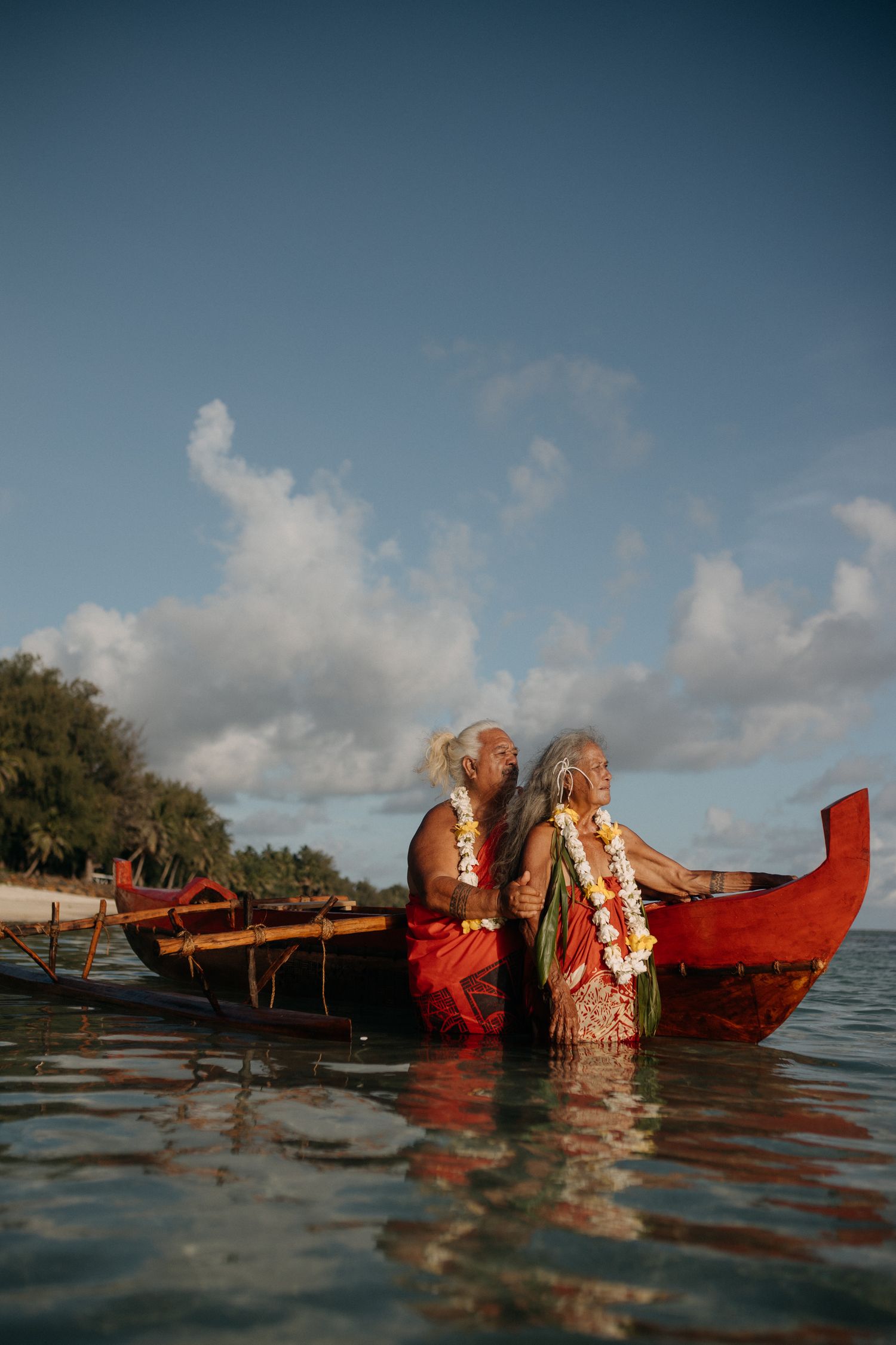 Two elderly Hawaiian elders in traditional leis and red garments sit back-to-back in a red outrigger canoe on calm waters.