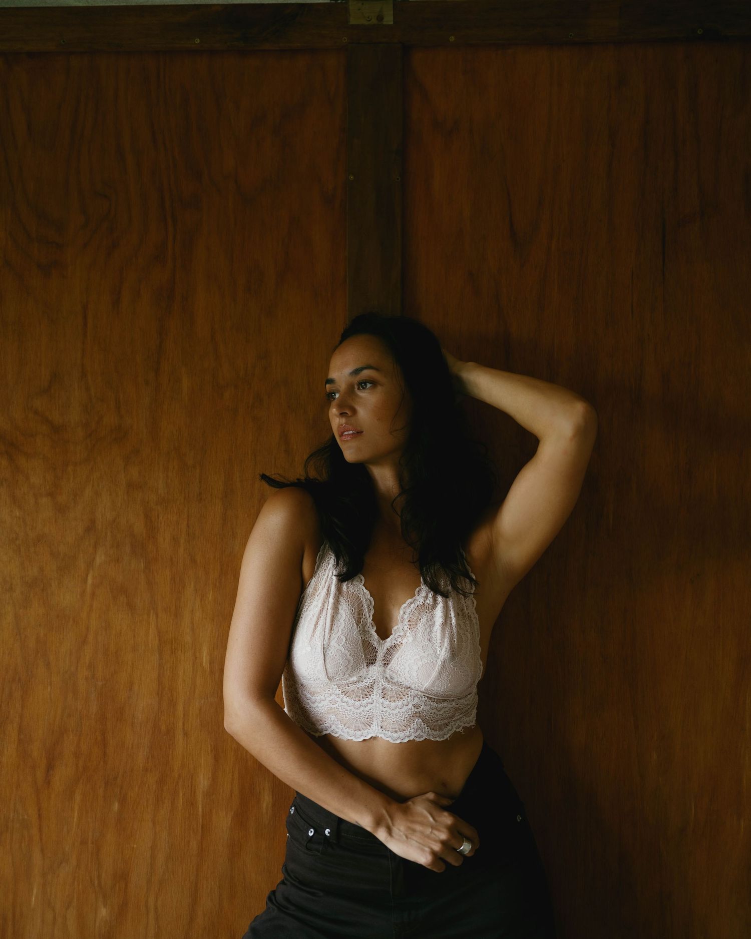 A young woman in a white lace crop top poses against a wooden wall, hand in hair, in moody natural lighting.