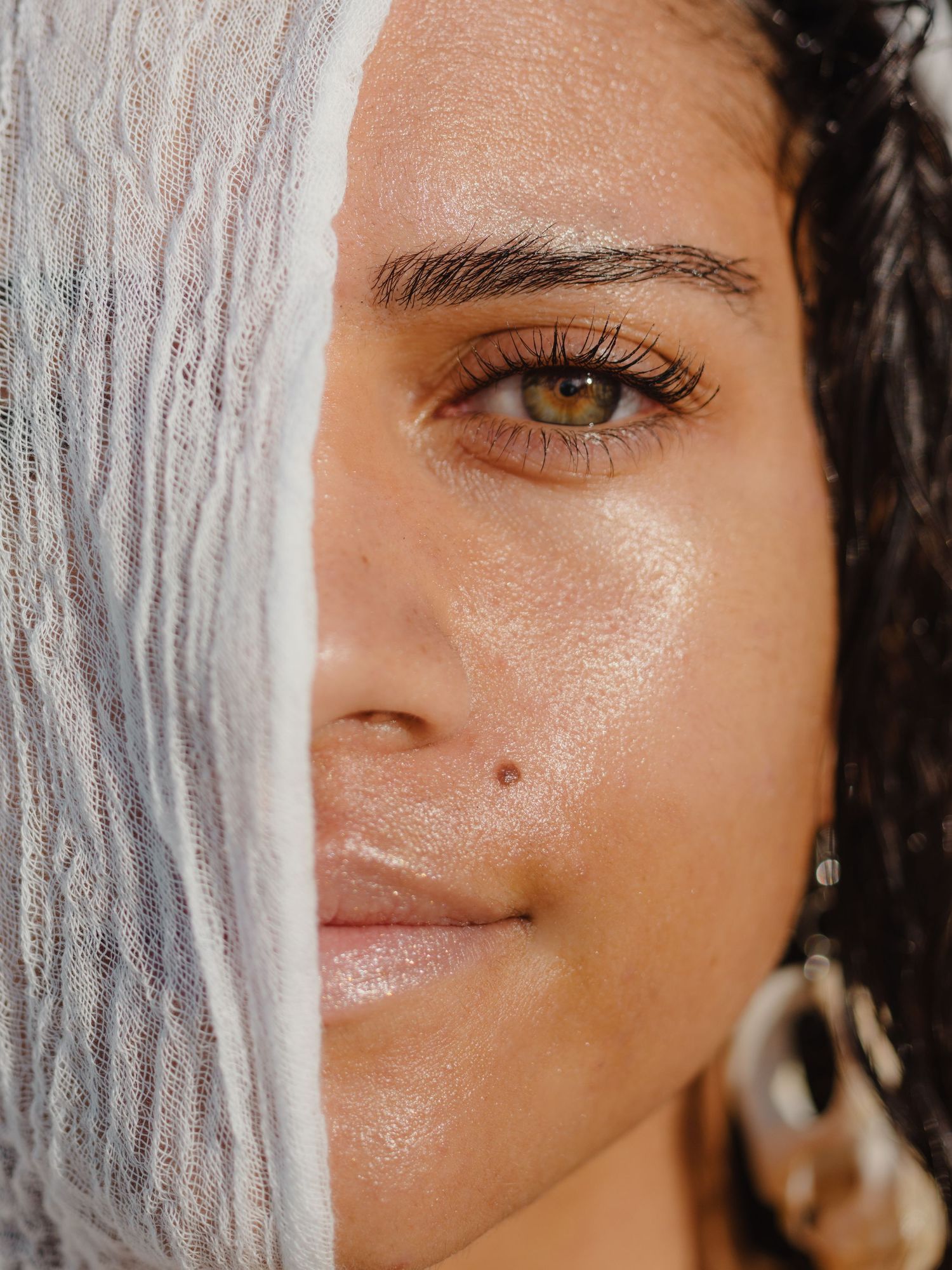 Close-up portrait of a woman with glowing skin, green eyes, and dark curly hair, half-covered by a white fabric.