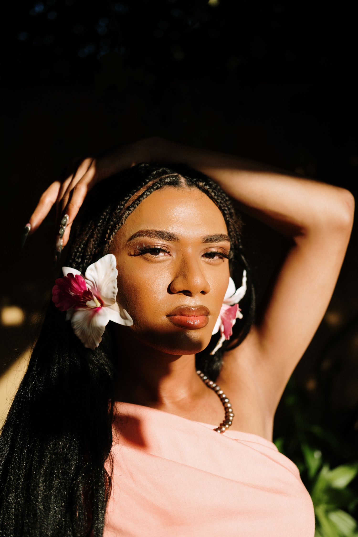 Young Black woman with braids and flowers in hair poses in sunlight wearing a pink top, with hands raised behind her head.