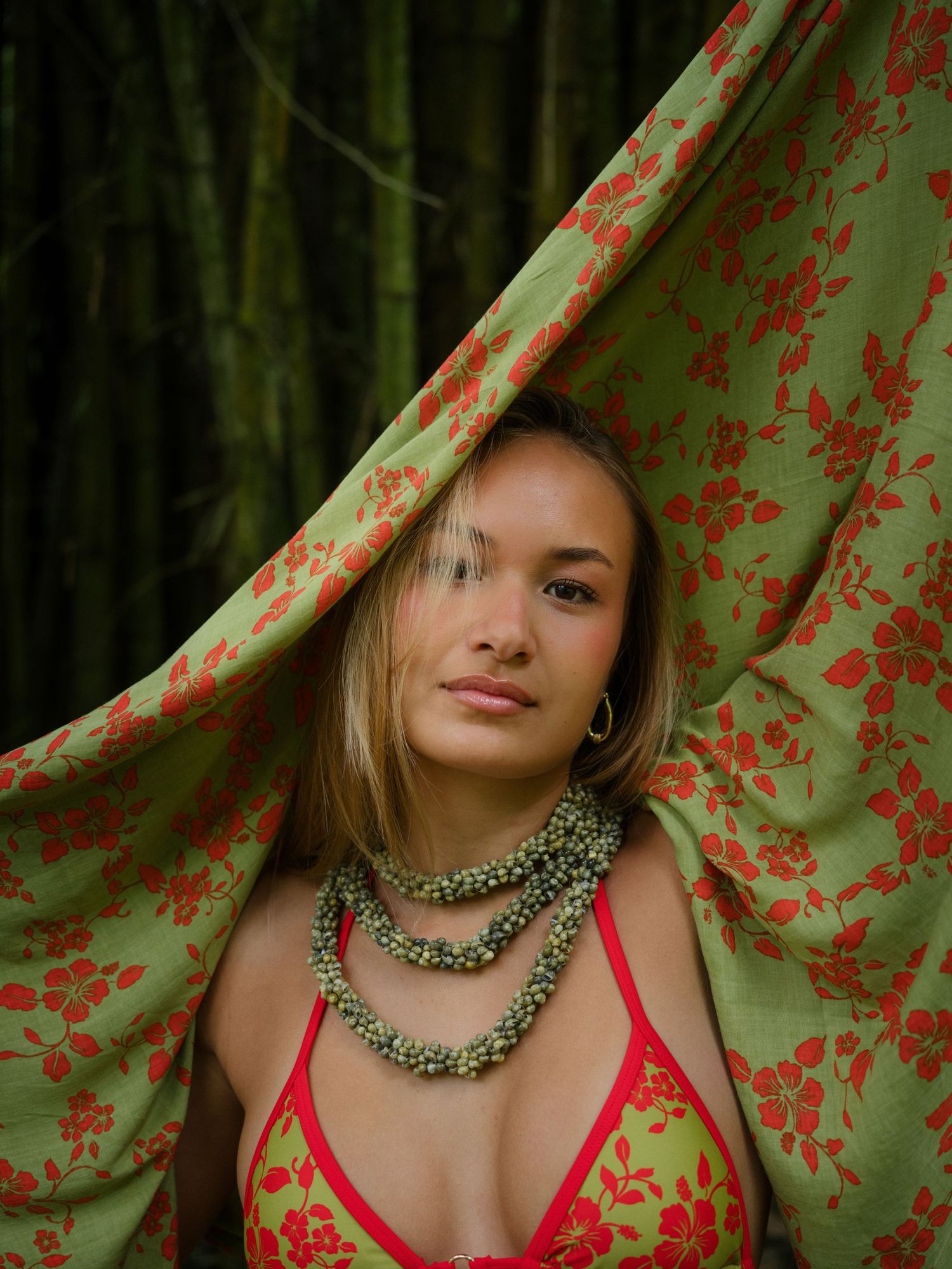 Young woman in red floral bikini and layered pearl necklace poses behind green red floral fabric with bamboo background.