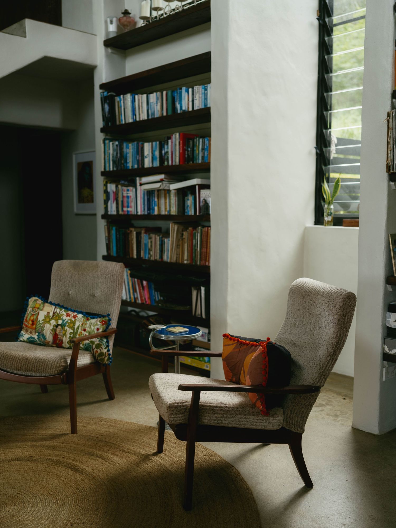 Cozy reading nook with two mid-century armchairs, colorful pillows, and a floor-to-ceiling bookshelf filled with books.
