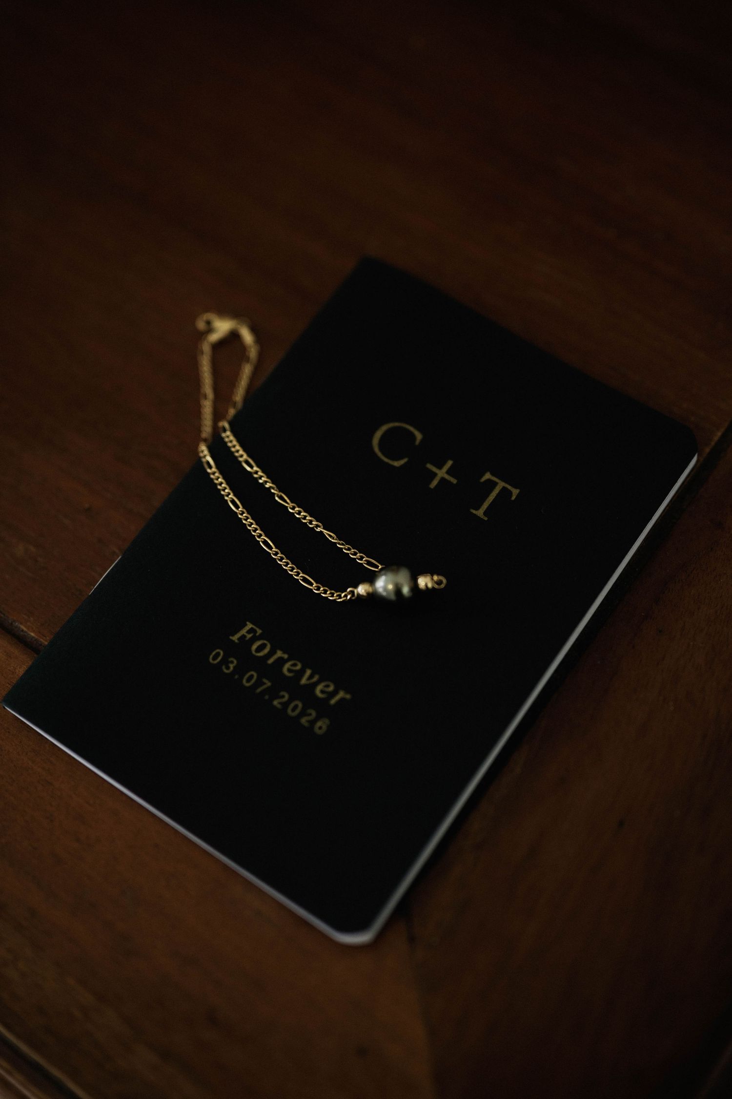Gold chain bracelet with pearl charm resting on black personalized wedding keepsake book engraved with C+T Forever.