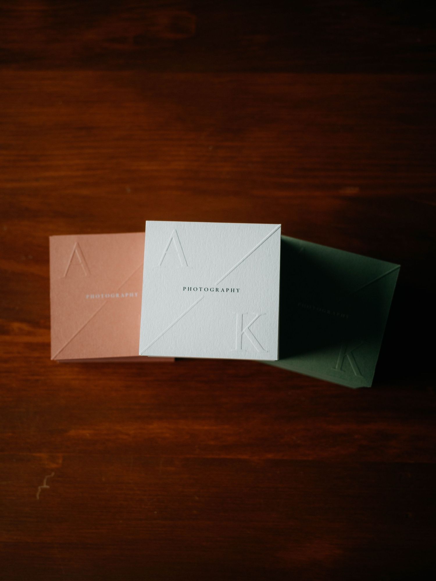 Three square photography brand boxes in pink, white, and green with embossed AK logo on dark wood surface.