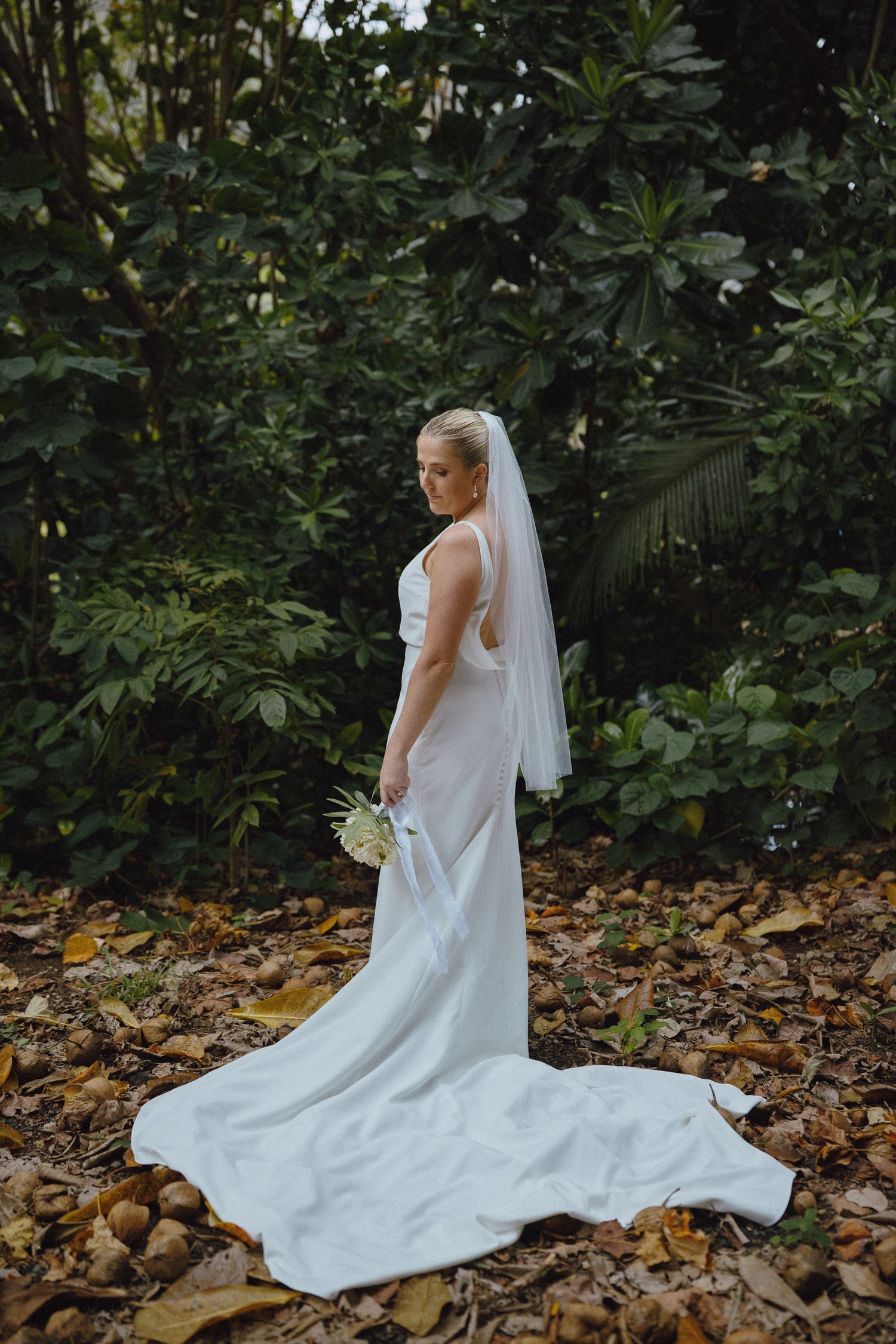 Bride in white gown and veil holding bouquet stands among autumn leaves before lush green tropical foliage.