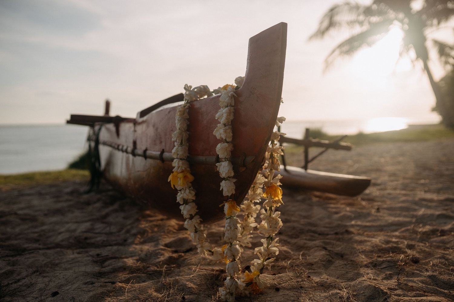 Traditional Hawaiian outrigger canoe on sandy beach at sunset with floral leis draped over bow near ocean.