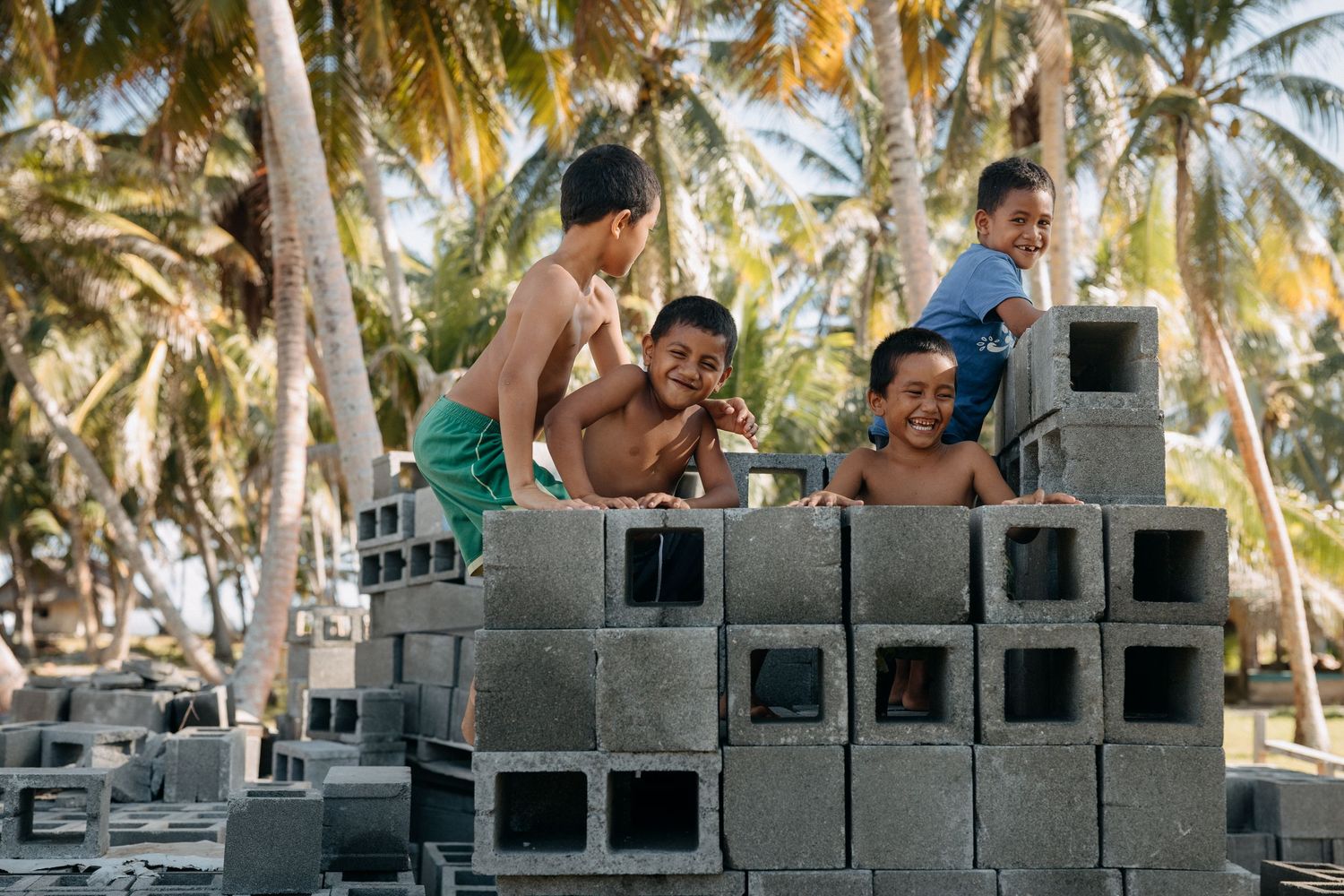 Children playing and stacking gray cinder blocks outdoors among tall palm trees in a tropical setting.