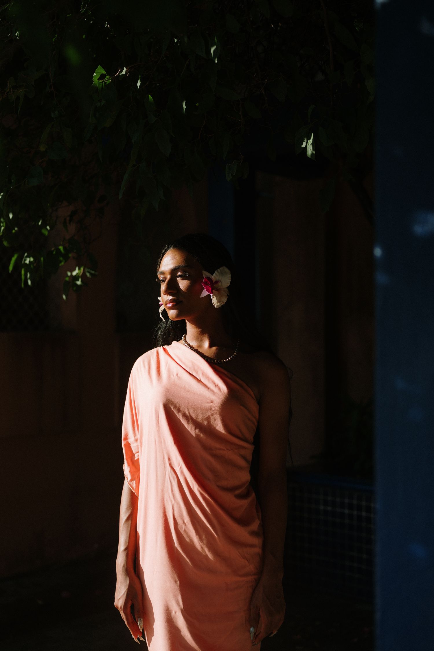 Woman in peach saree with flower in hair stands in dramatic shadow and light outdoors.