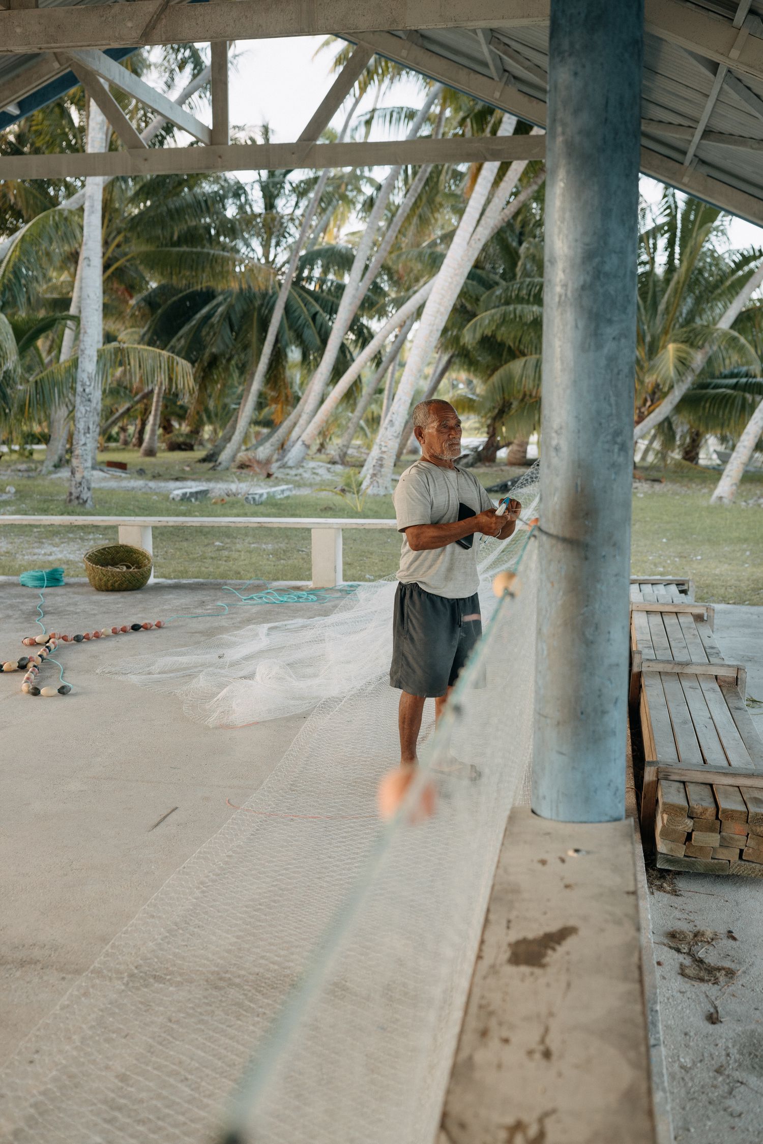 A man repairs a fishing net under an open-air shelter surrounded by tropical palm trees.