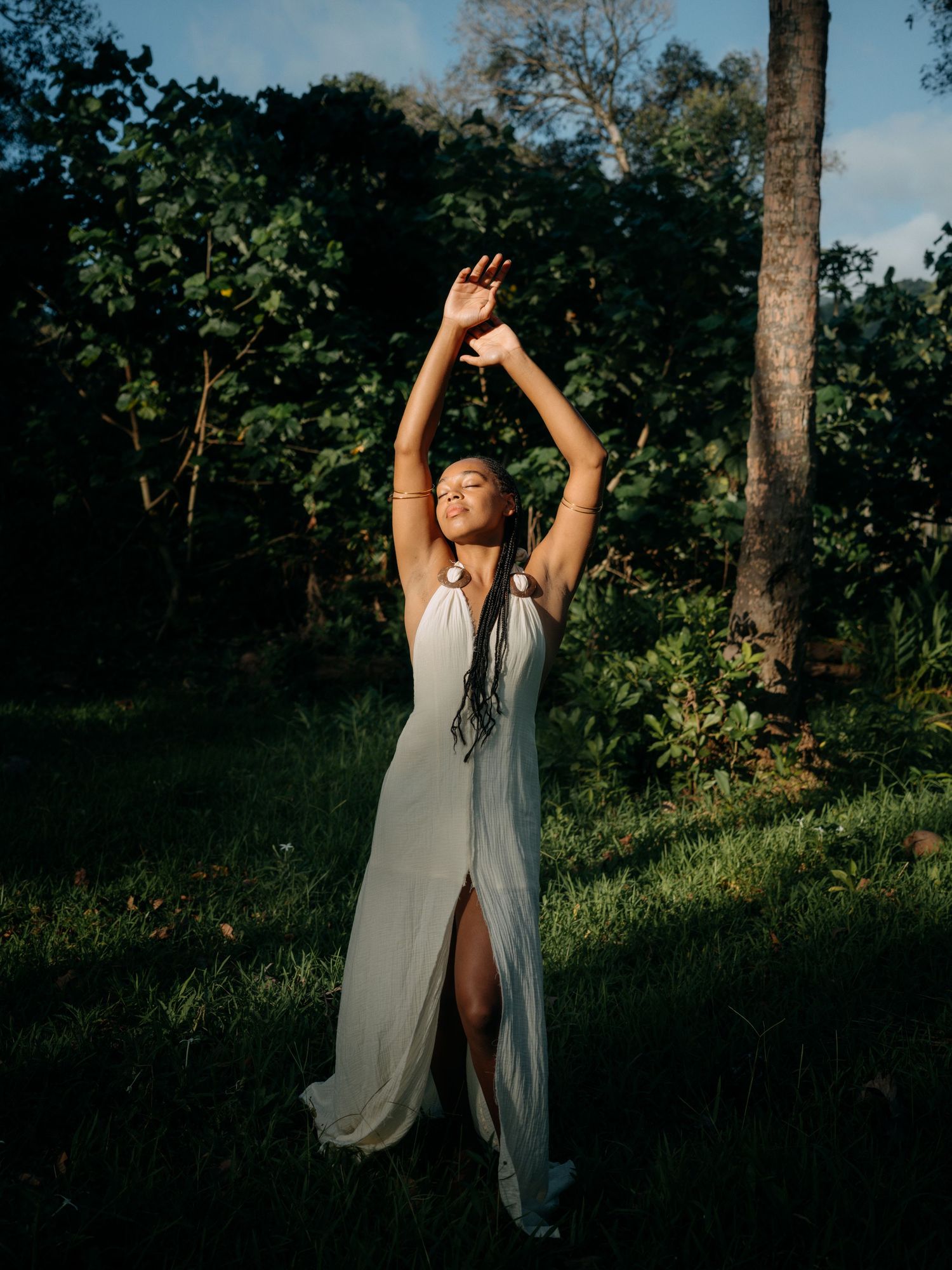 Woman in white halter maxi dress with slit poses gracefully outdoors, arms raised, surrounded by lush green trees.