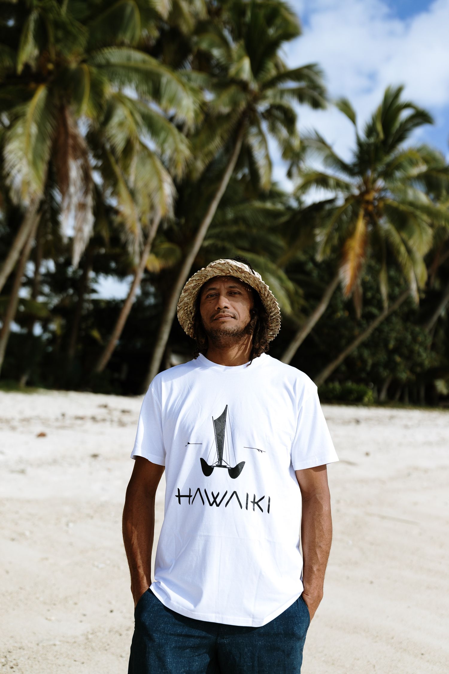 Man wearing Hawaiki white t-shirt and straw hat standing on tropical beach with palm trees.