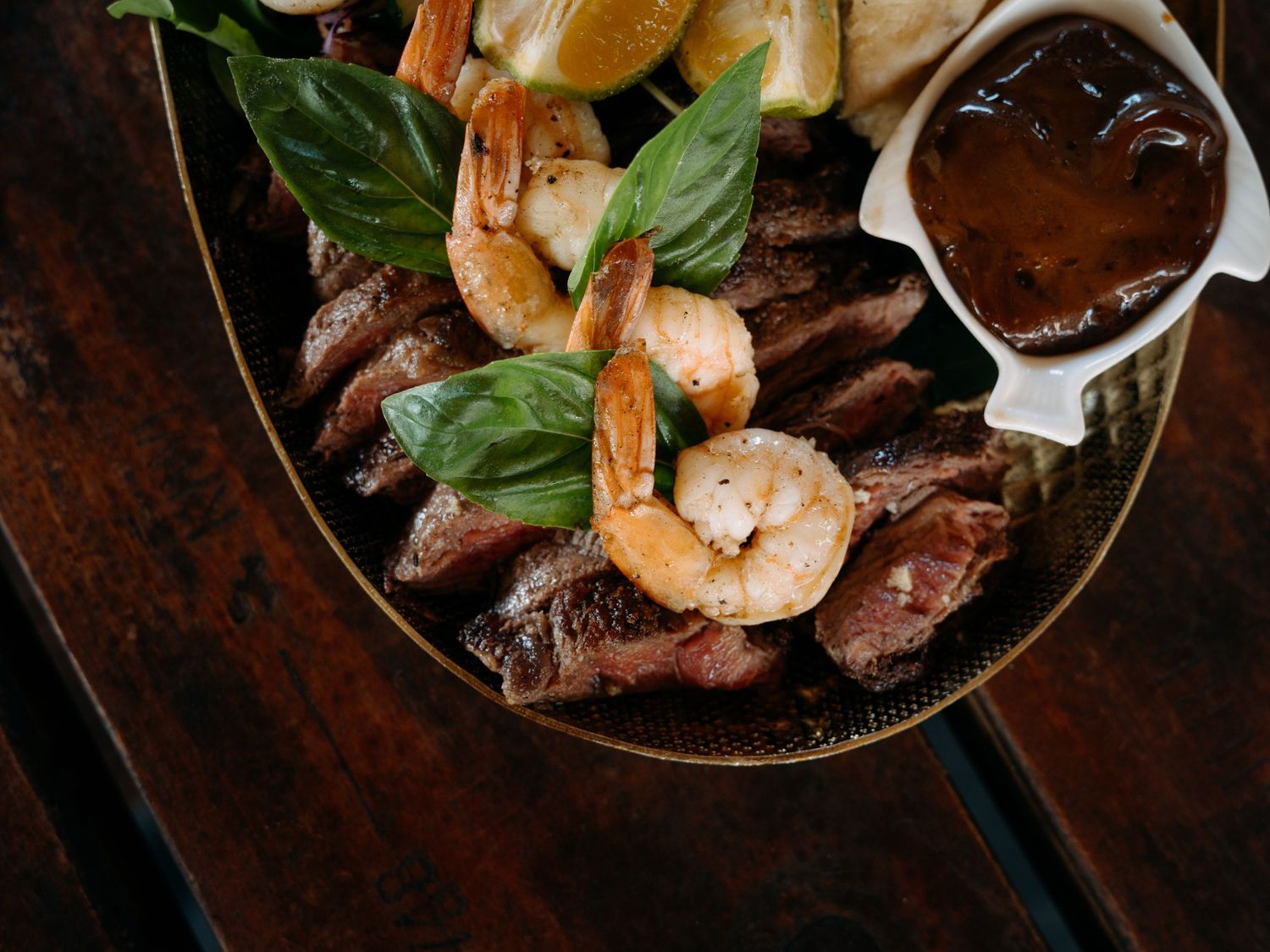 Surf and turf dish with grilled shrimp, sliced steak, fresh basil, and BBQ dipping sauce served in a bowl.