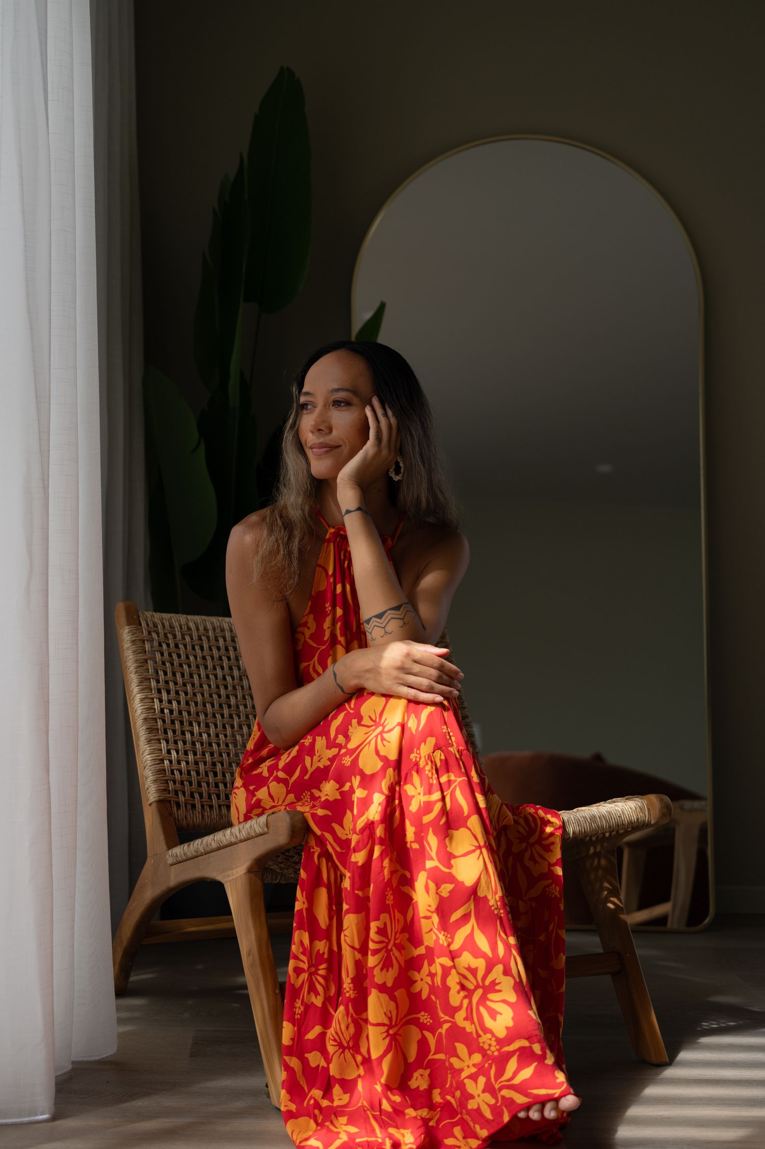 Woman in red floral halter maxi dress seated in rattan chair by mirror and tropical plant.