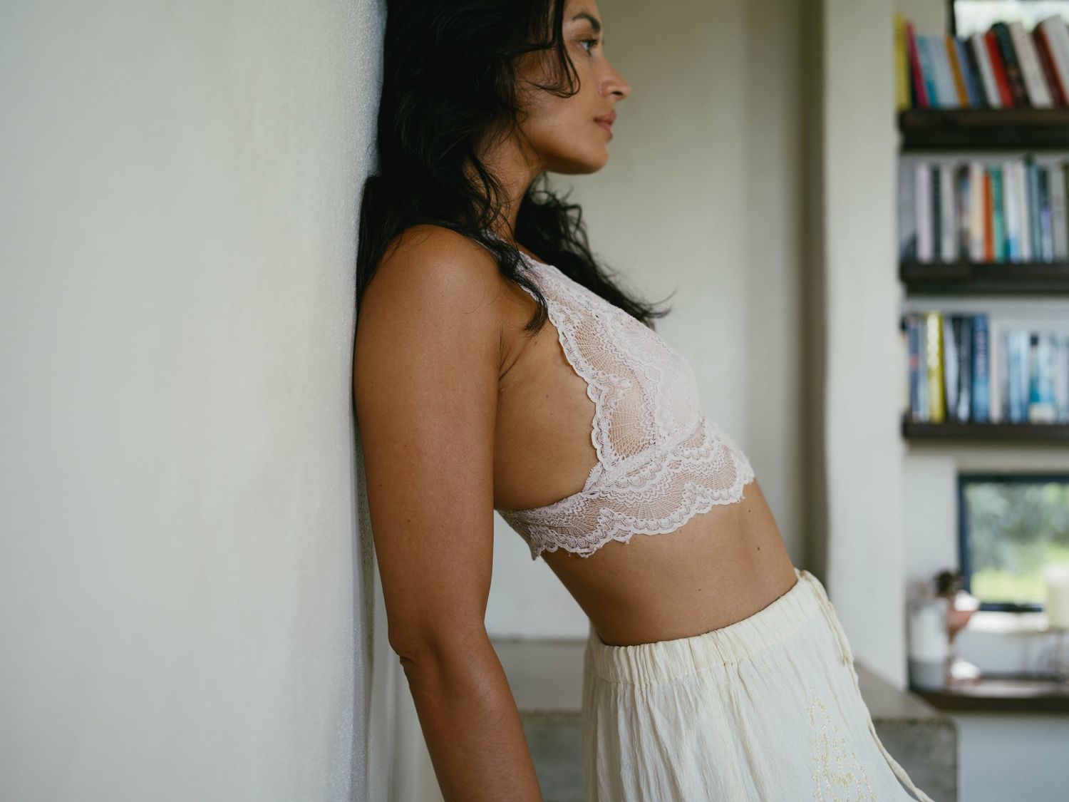 Woman in white lace bralette and flowing skirt leaning against a wall in a serene, softly lit room.