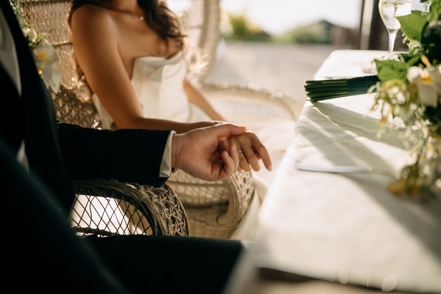 Bride and groom holding hands at wedding table with flowers, romantic ceremony moment in warm sunlight.