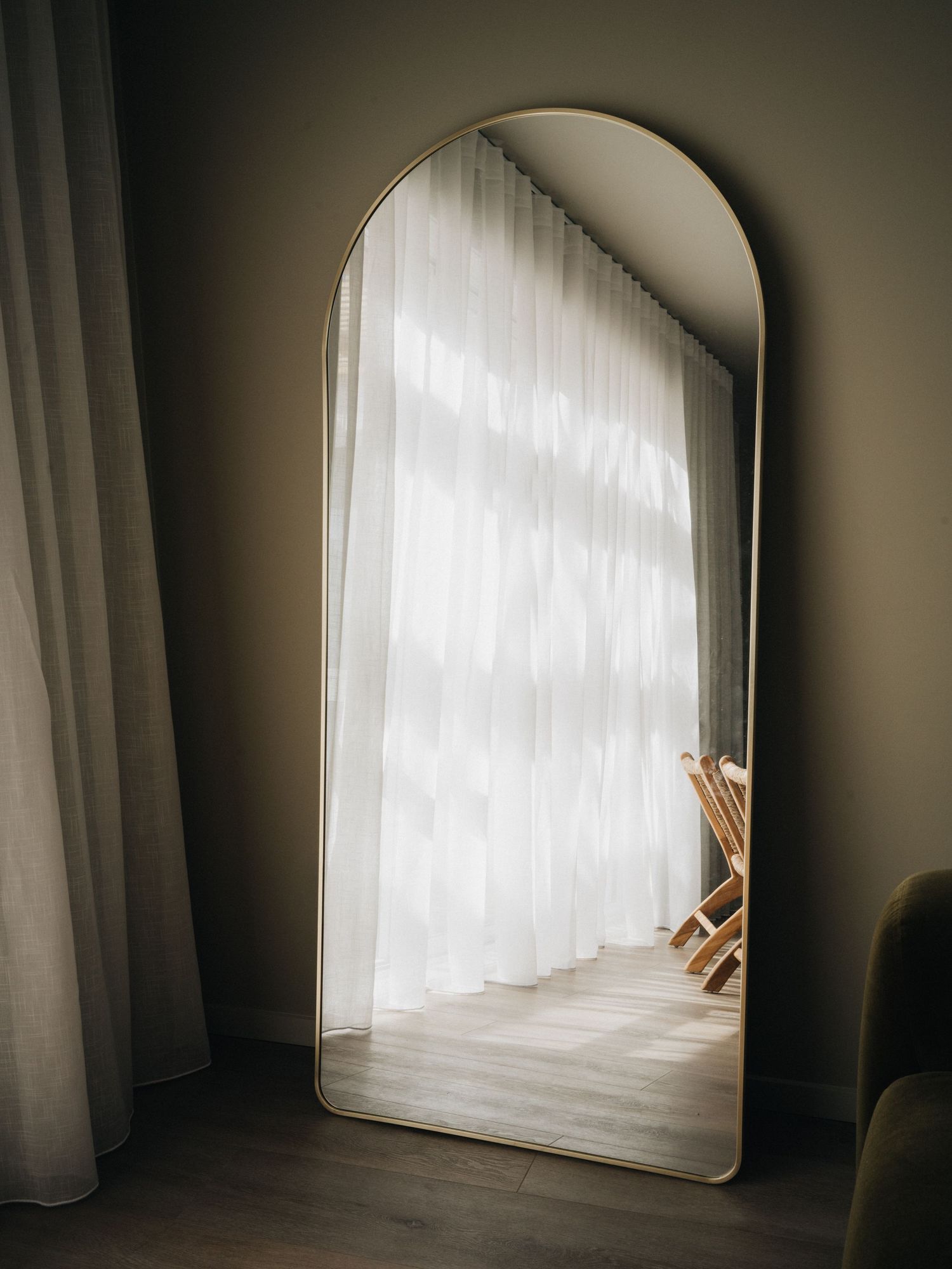 Tall arched full-length floor mirror with gold frame leaning against wall near sheer white curtained window.