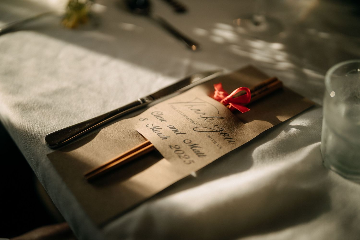 Elegant wedding place setting with calligraphy menu card, chopsticks, and red bow on white linen tablecloth.