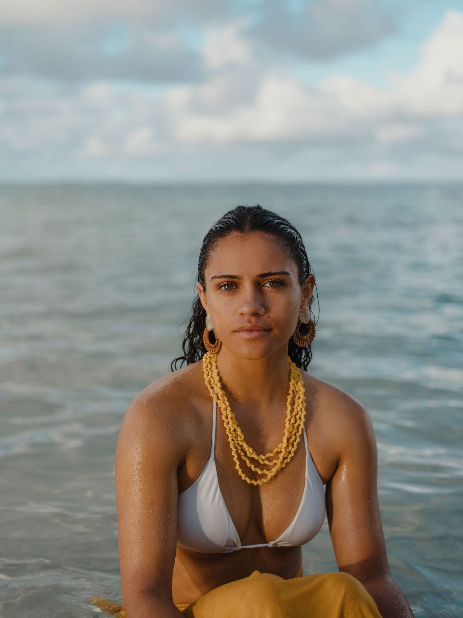 Woman in white bikini top and gold necklaces sitting by the ocean on a cloudy day.