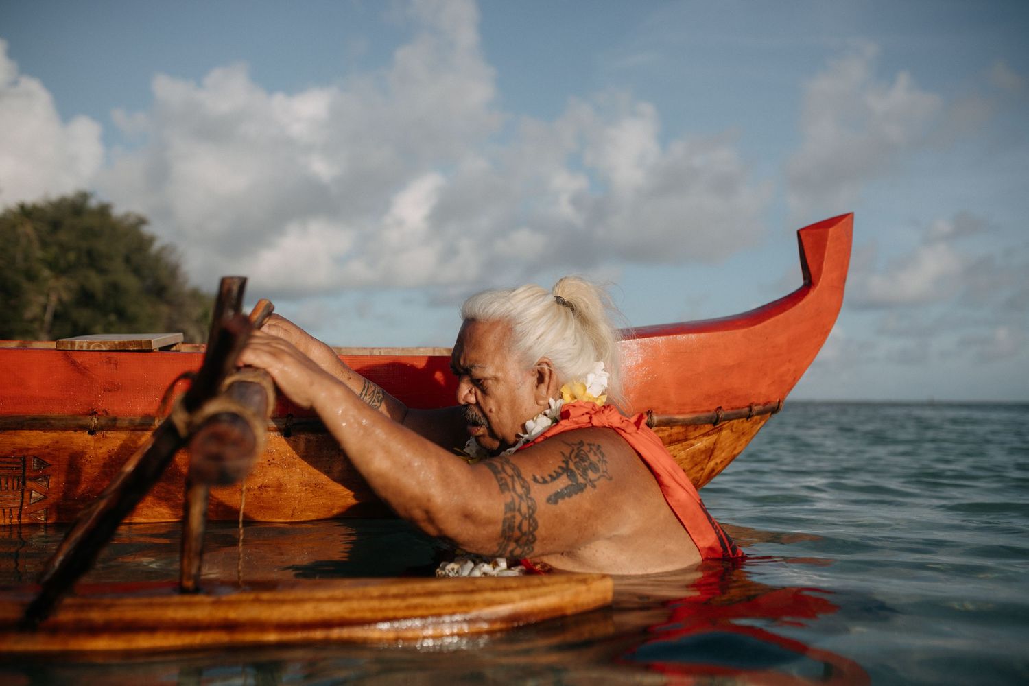 Elderly tattooed man paddling a red outrigger canoe on calm ocean waters under a partly cloudy sky.